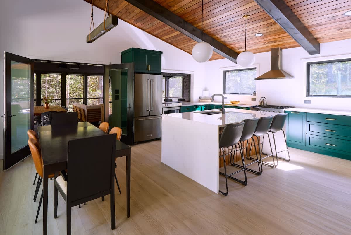 An open-concept kitchen and dining area features a vaulted wood-paneled ceiling with dark beams, a large white island with bar seating, and dark green cabinetry. A dining table with chairs is visible near large glass doors leading to an outdoor space.