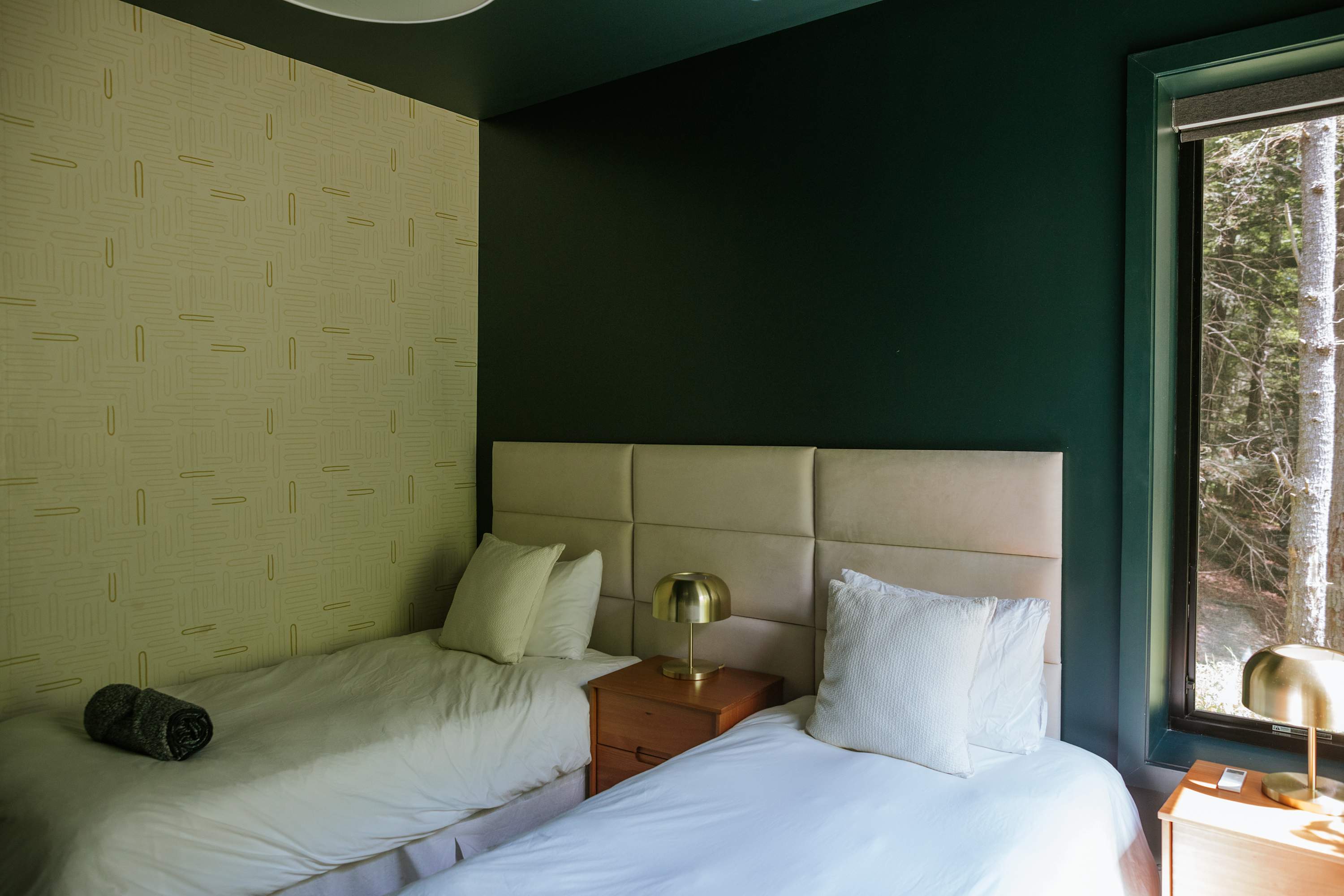 A different angle of a bedroom shows two twin beds with white bedding and upholstered headboards, one against a dark green wall and the other against a light green patterned wallpaper, with a wooden nightstand and gold lamp between them.