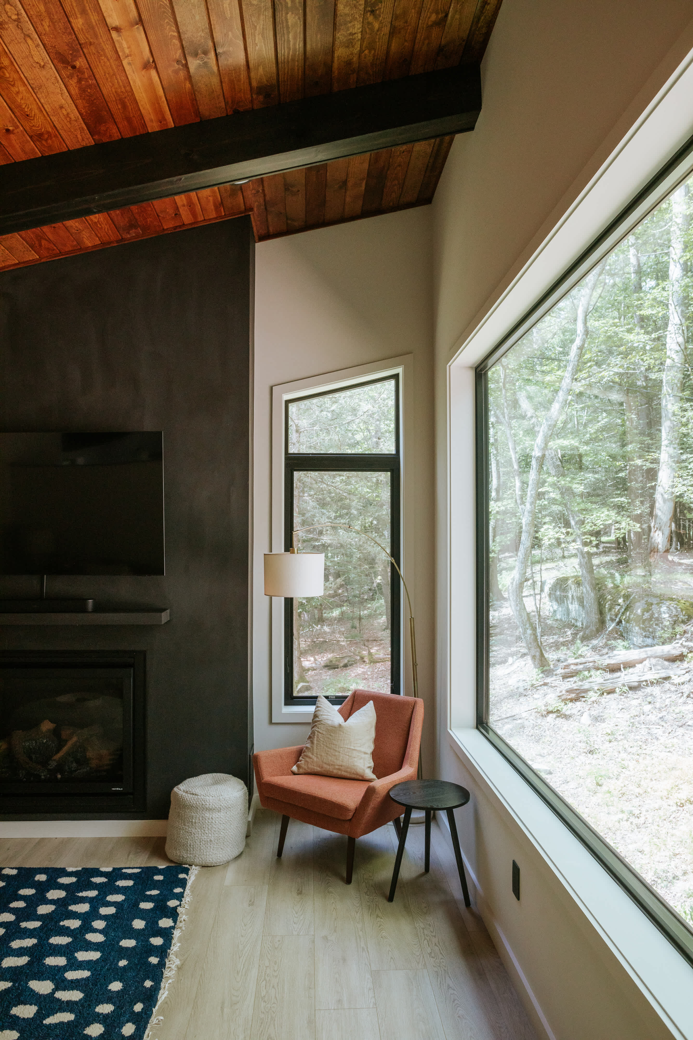A bright living room features a modern fireplace with a dark textured surround and a mounted TV, alongside an orange accent chair and a floor lamp. Large windows offer expansive views of the surrounding forest, complementing the wood-paneled ceiling.