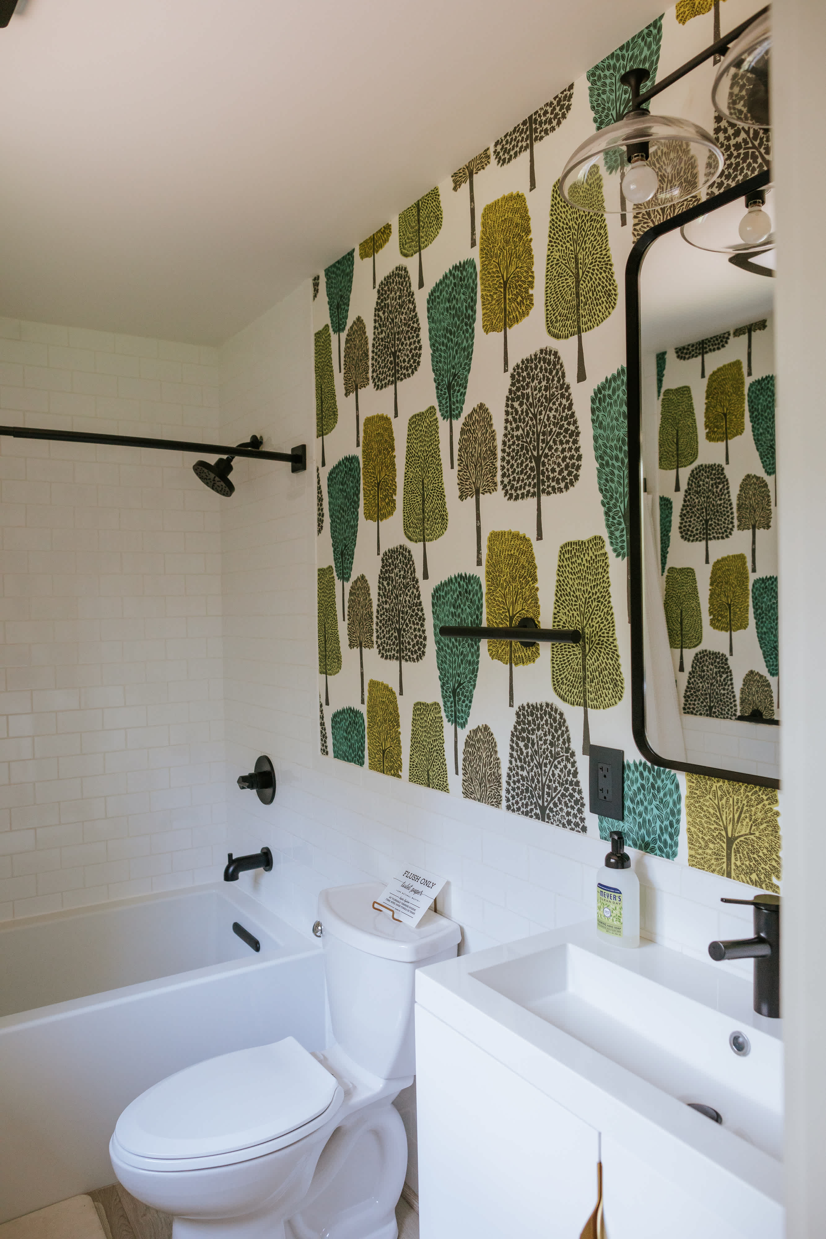 A bright bathroom features a white subway tile shower/tub combo and a white vanity with a black faucet, complemented by a striking wallpaper with a pattern of green and yellow trees. A black-framed mirror and black fixtures add modern accents.