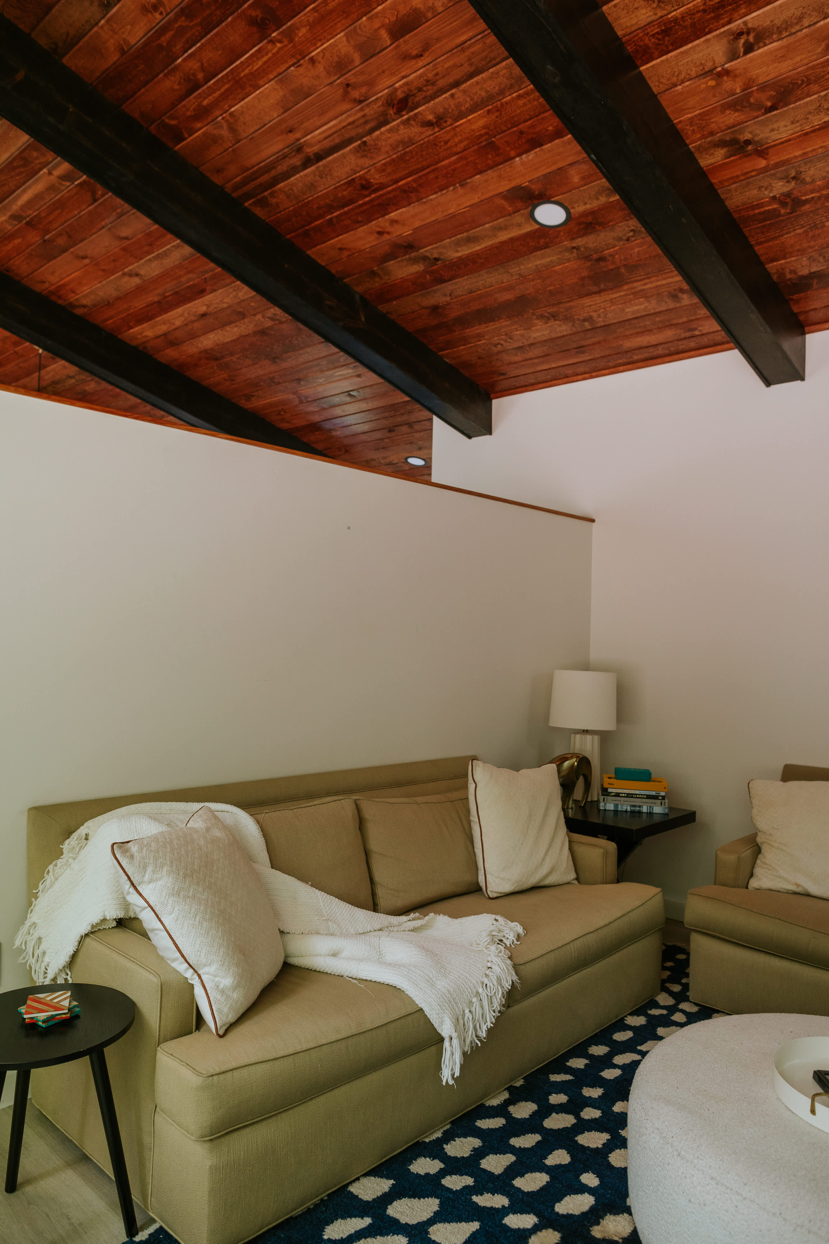 A cozy living area features a light tan sofa adorned with white throw pillows and a blanket, positioned on a dark blue rug with white polka dots. The room has a warm wood-paneled ceiling with dark exposed beams and a small side table.