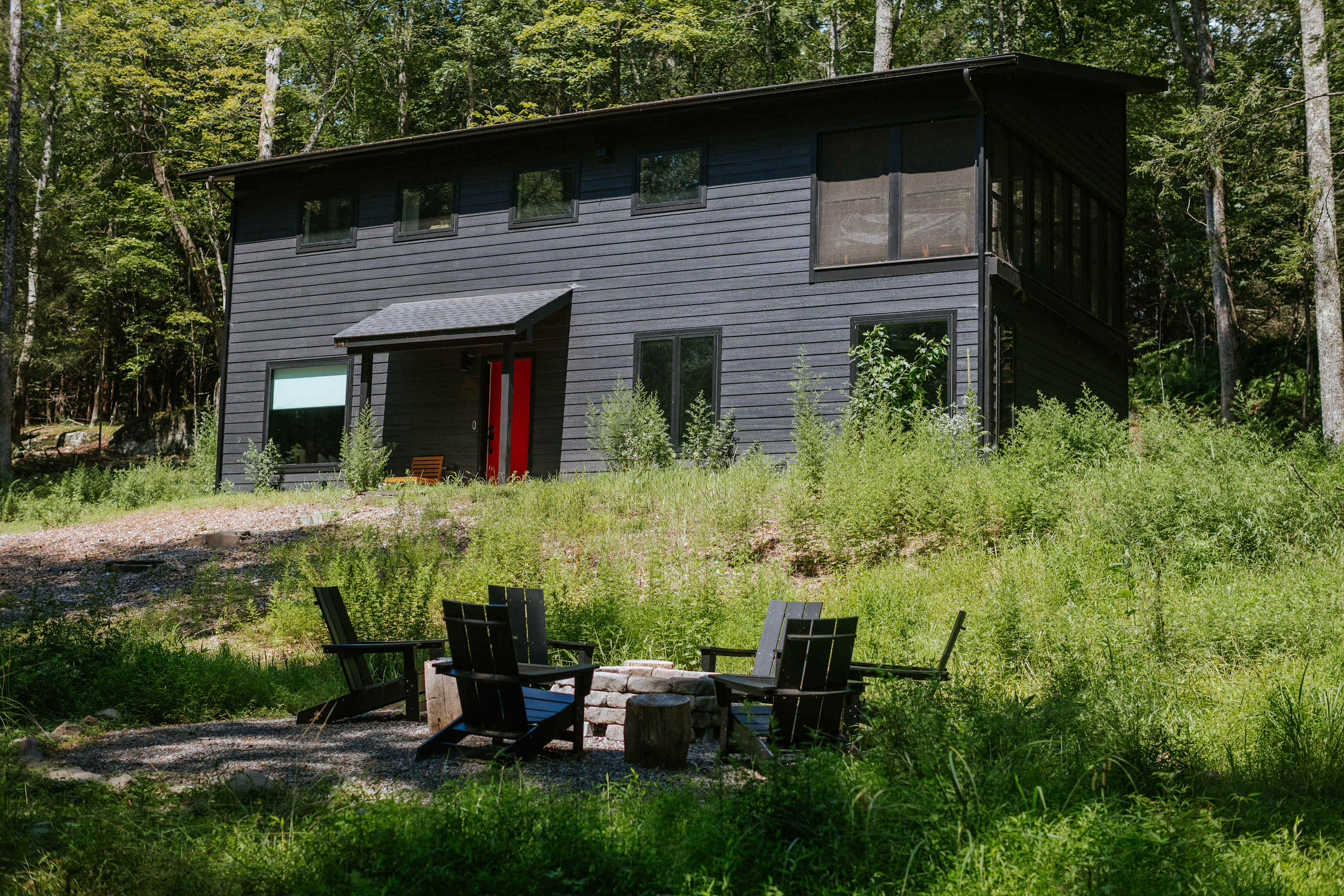 Modern black house with a red front door, surrounded by lush green foliage, featuring a gravel fire pit area with Adirondack chairs in the foreground.