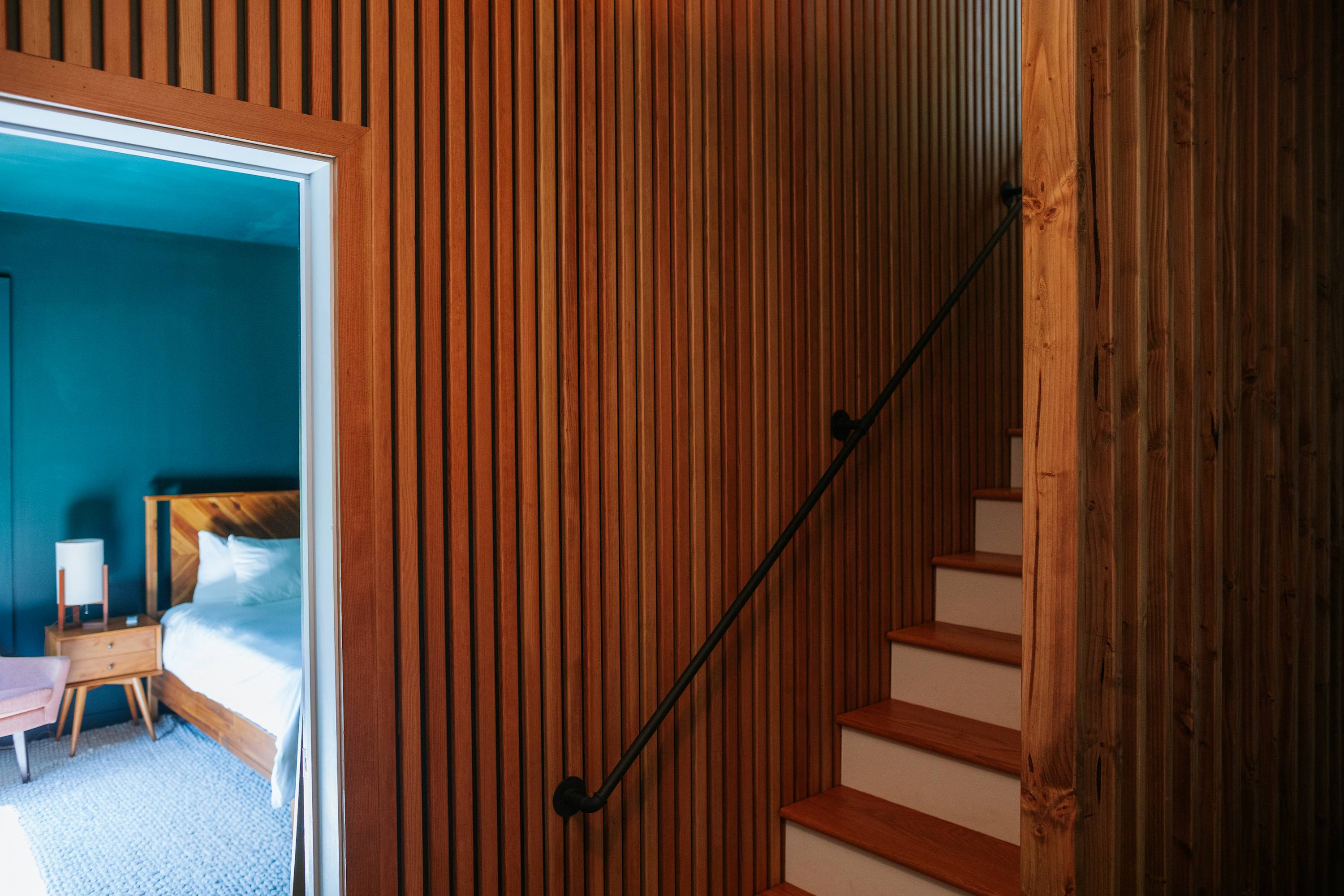 A modern bedroom with dark teal walls and a wooden bed frame is visible through an open doorway, adjacent to a staircase featuring warm wooden slat walls and a black metal handrail.