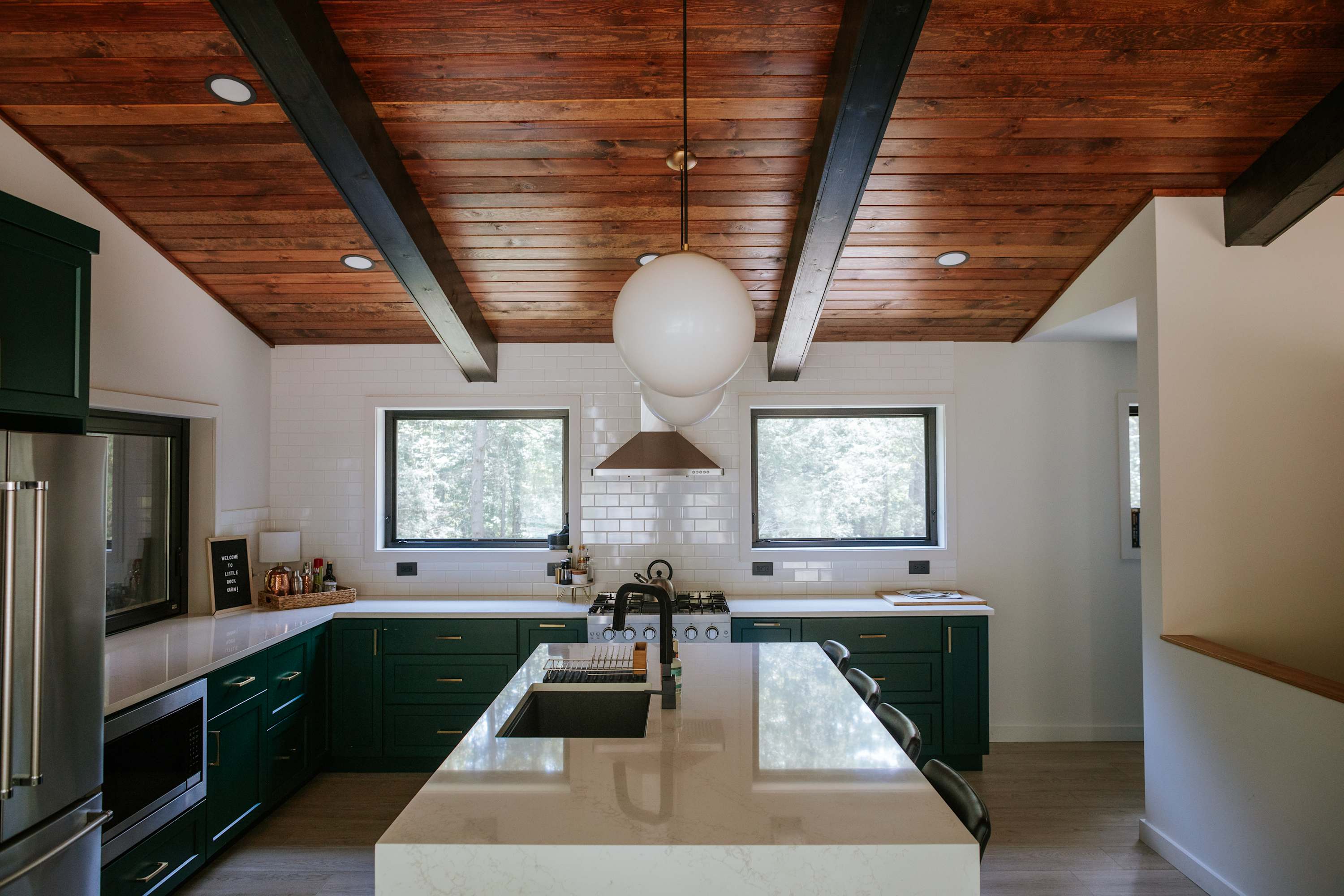 A modern kitchen showcases dark green lower cabinets, white subway tile backsplash, and a large white quartz island with an integrated sink and black faucet. The vaulted wood-paneled ceiling with dark beams adds warmth, complemented by two large pendant lights.