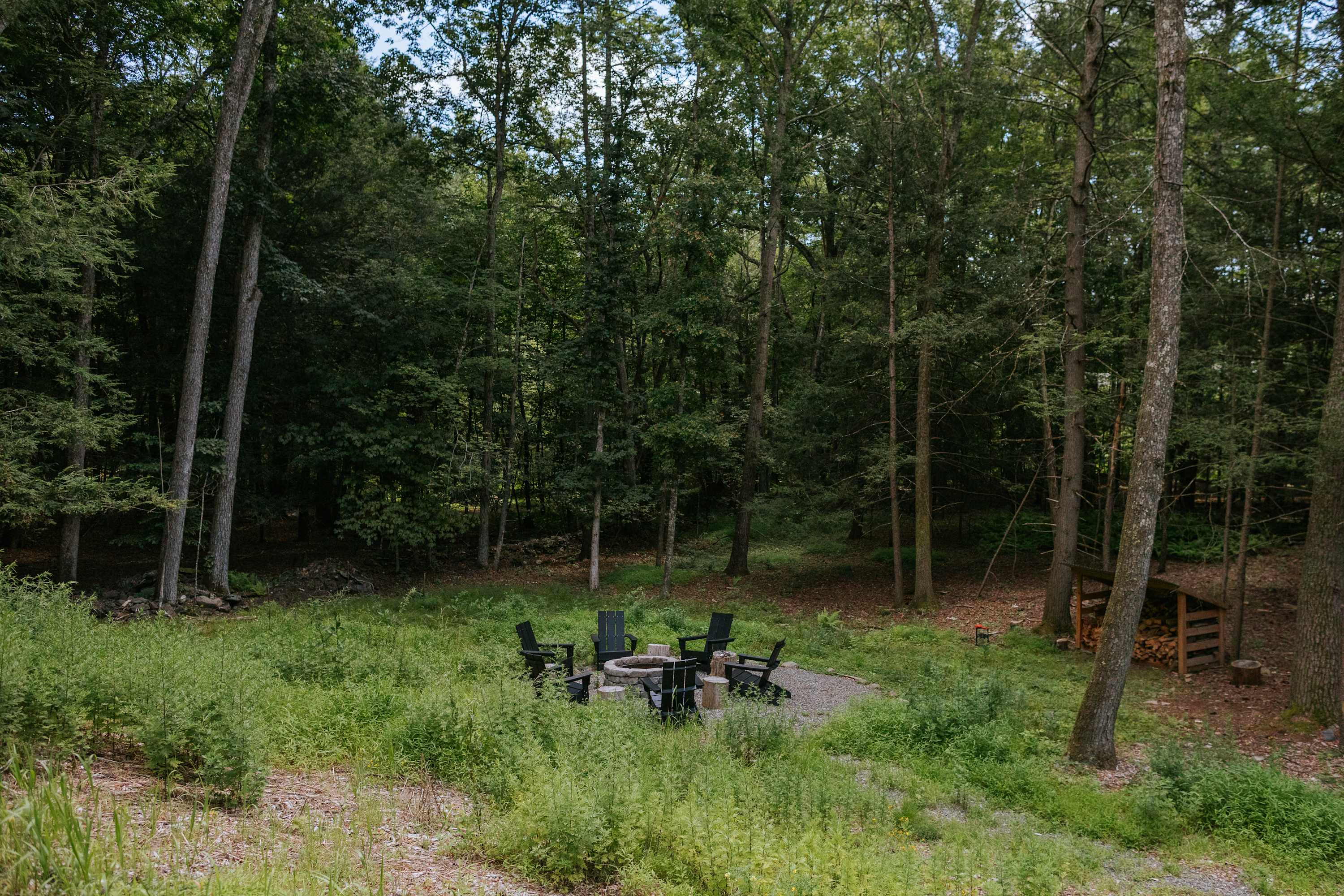 Outdoor fire pit area surrounded by four black Adirondack chairs, set on a gravel base in a grassy clearing within a dense forest. A stack of firewood is visible nearby, indicating a rustic outdoor gathering space.