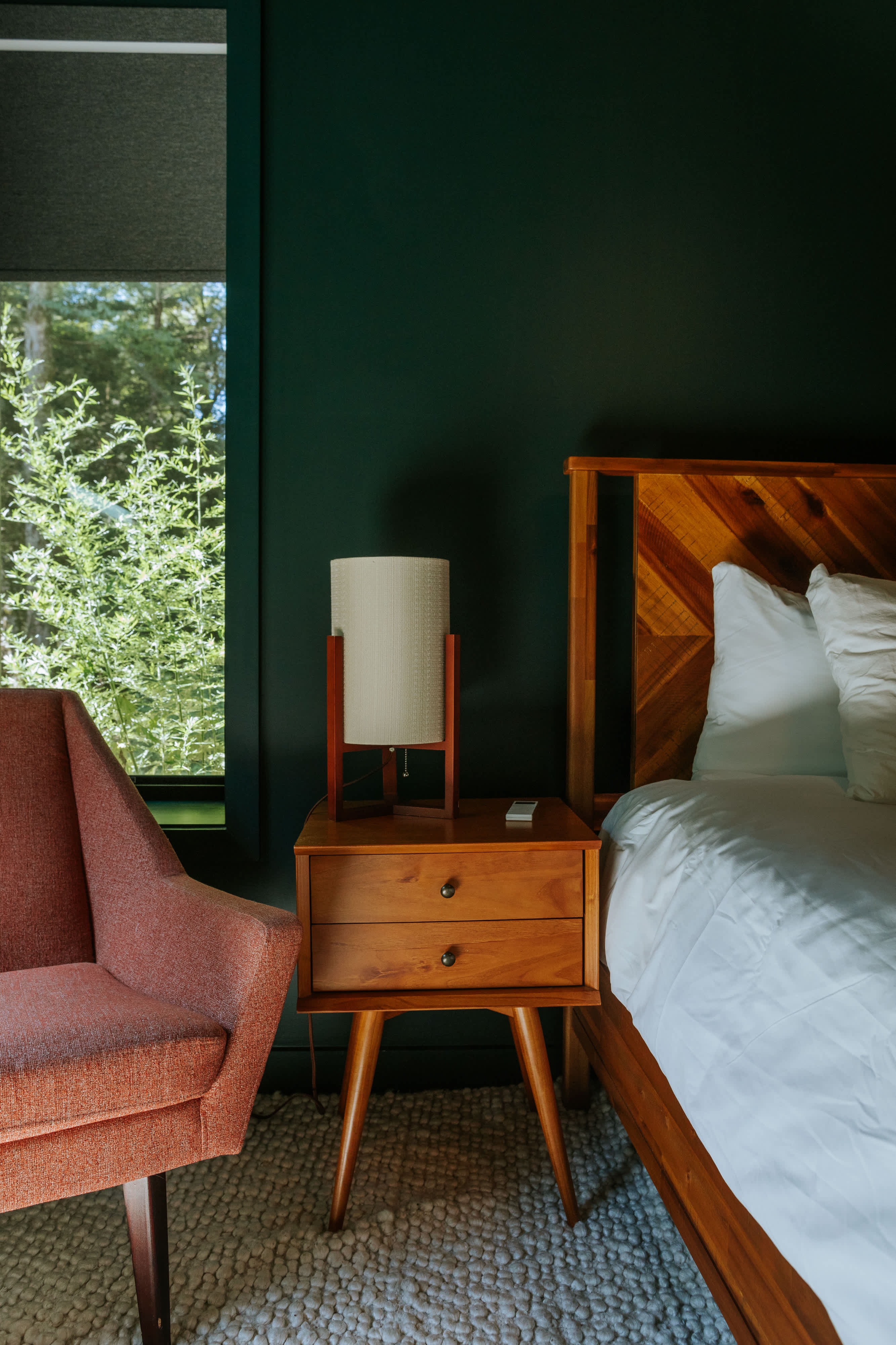A cozy bedroom with dark green walls features a wooden bed frame with a patterned headboard, white bedding, a mid-century modern nightstand with a lamp, and a coral-colored armchair, all on a textured cream rug.