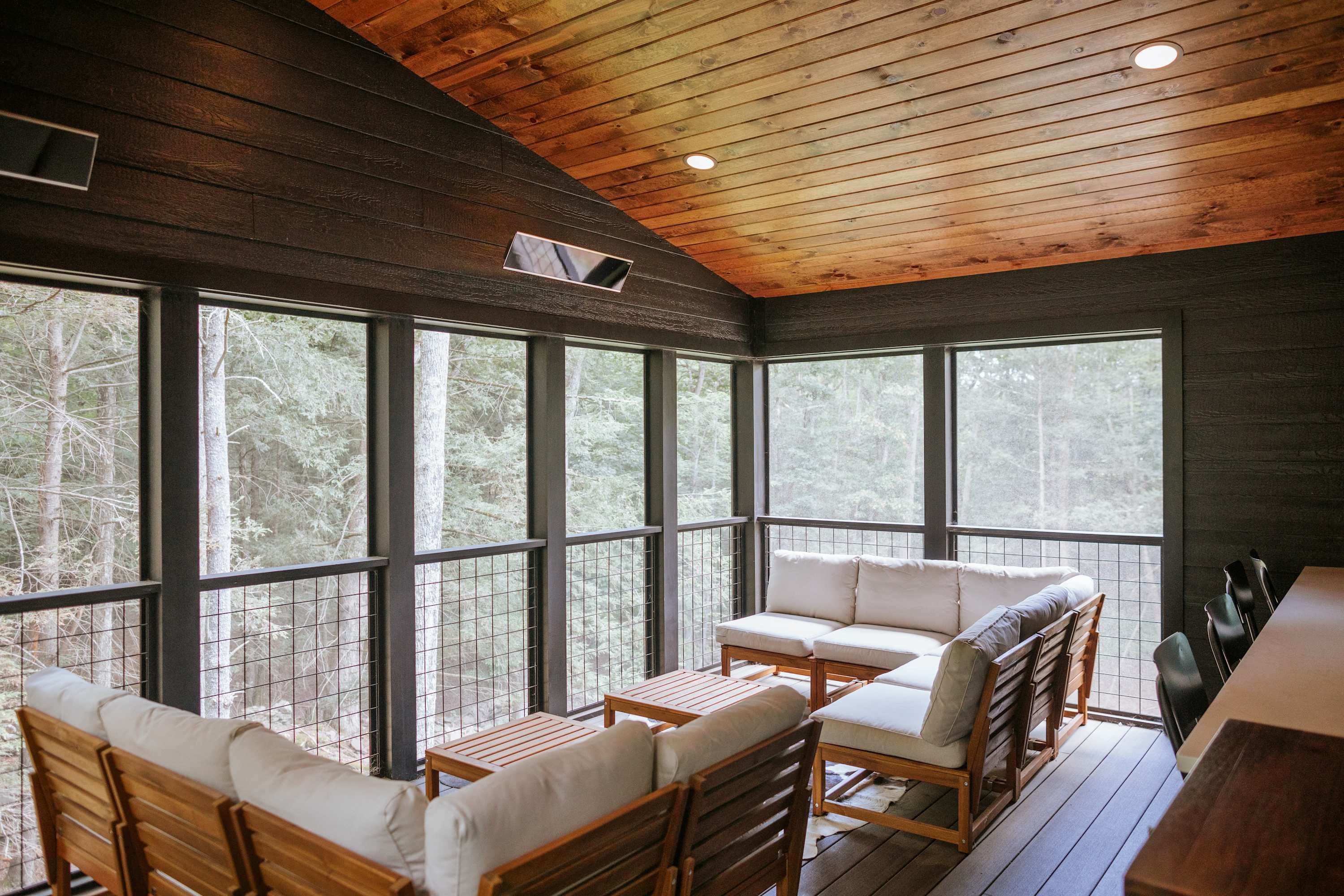 Enclosed screened porch with light wood outdoor seating featuring white cushions, offering panoramic views of the surrounding dense forest. The space has a warm wooden sloped ceiling with dark beams and recessed lighting.