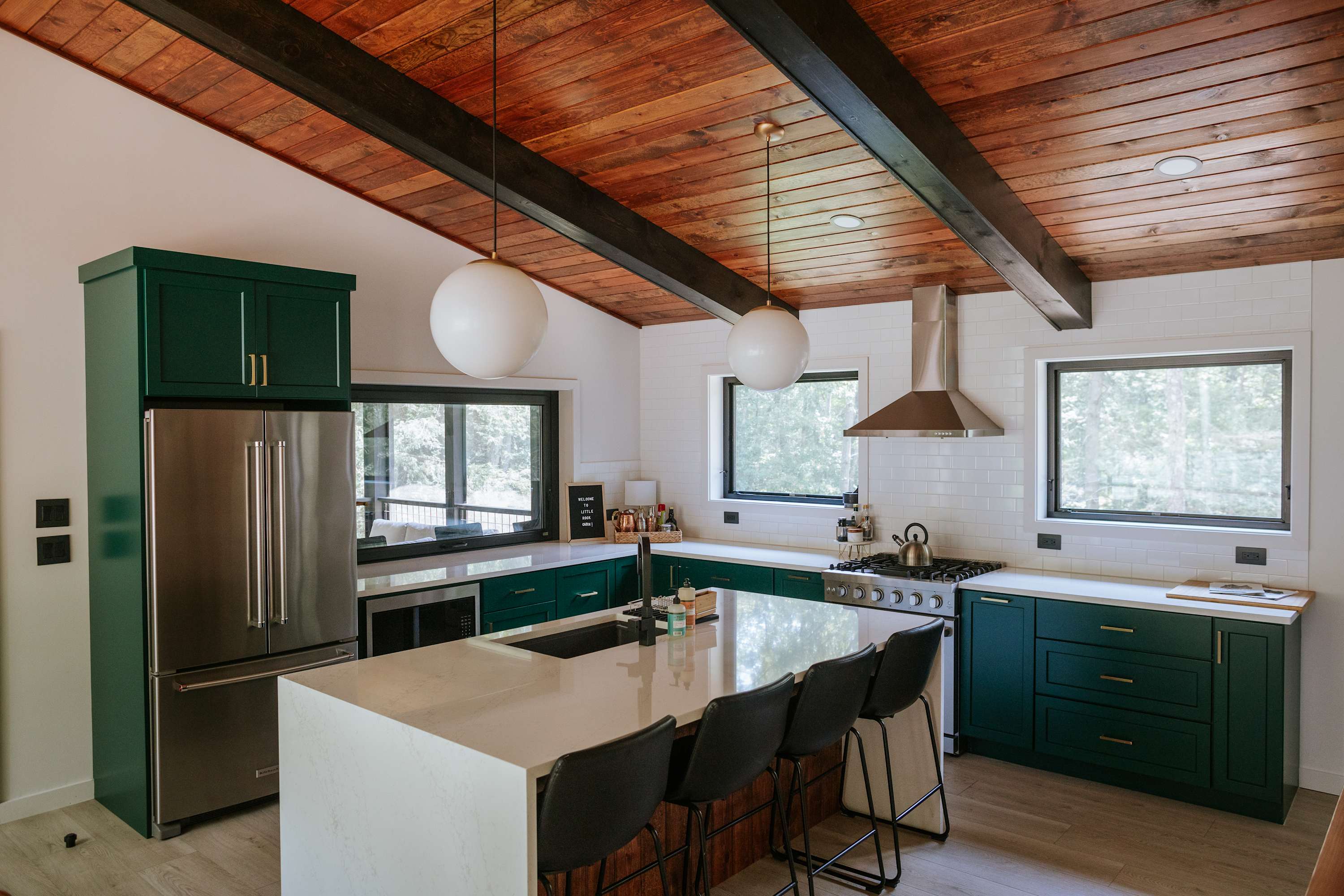 Spacious kitchen with dark green cabinets, white quartz countertops, and a large island with bar seating. Stainless steel appliances are integrated, and large windows overlook the wooded surroundings, all beneath a warm wooden sloped ceiling.