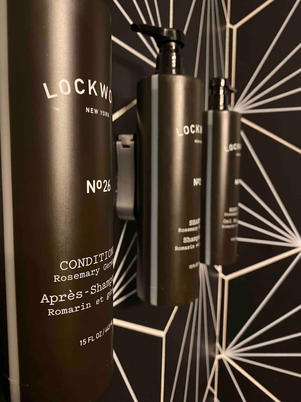 A close-up shot displays three dark-colored pump bottles of 'LOCKWOOD NEW YORK' brand conditioner, shampoo, and body wash, mounted on a wall with a distinctive black and white geometric pattern.