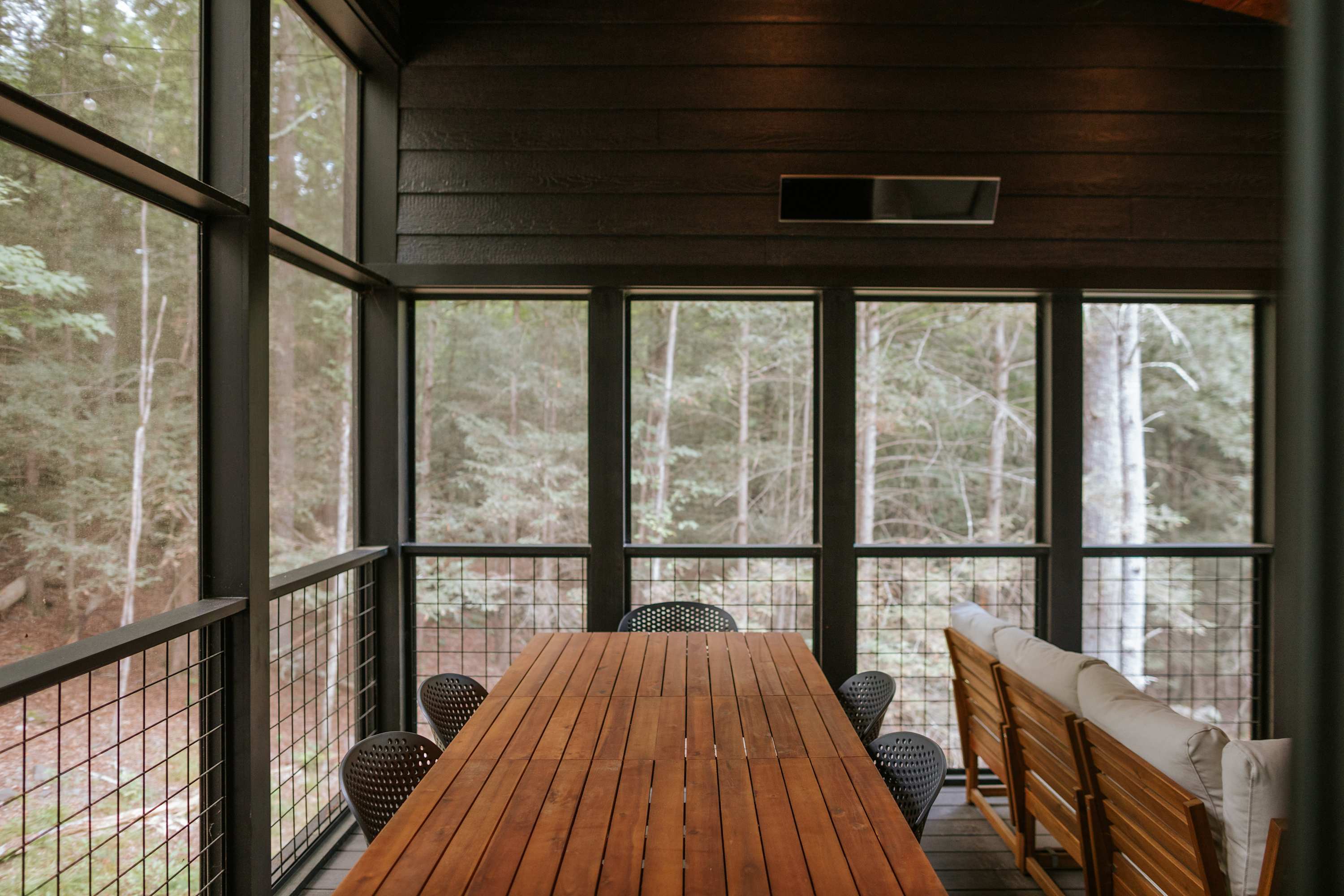 A spacious screened porch features a long wooden dining table with black chairs, and a light-colored outdoor sofa, all surrounded by large windows offering immersive views of the dense forest. The dark wood walls and ceiling create a cozy outdoor living space.