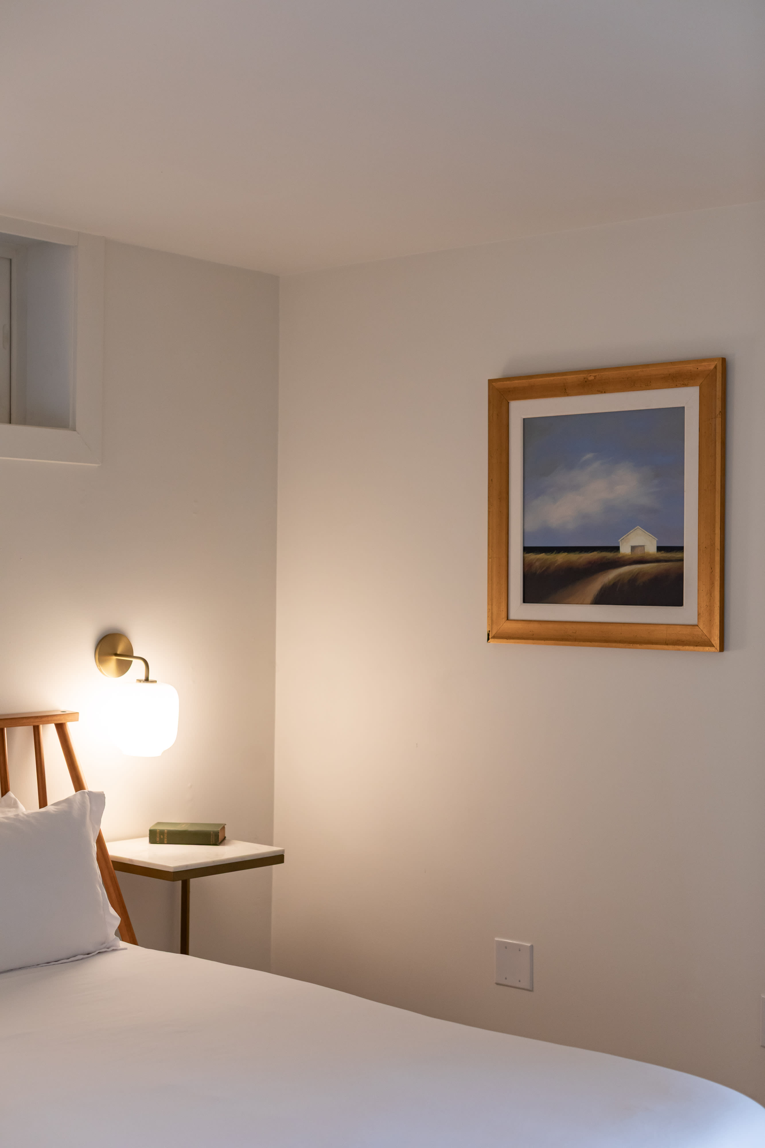 A close-up of the wooden bed frame with white bedding and a side table, featuring a brass wall sconce and a book. A framed painting of a white house in a field hangs on the wall, adding a touch of art to the space.