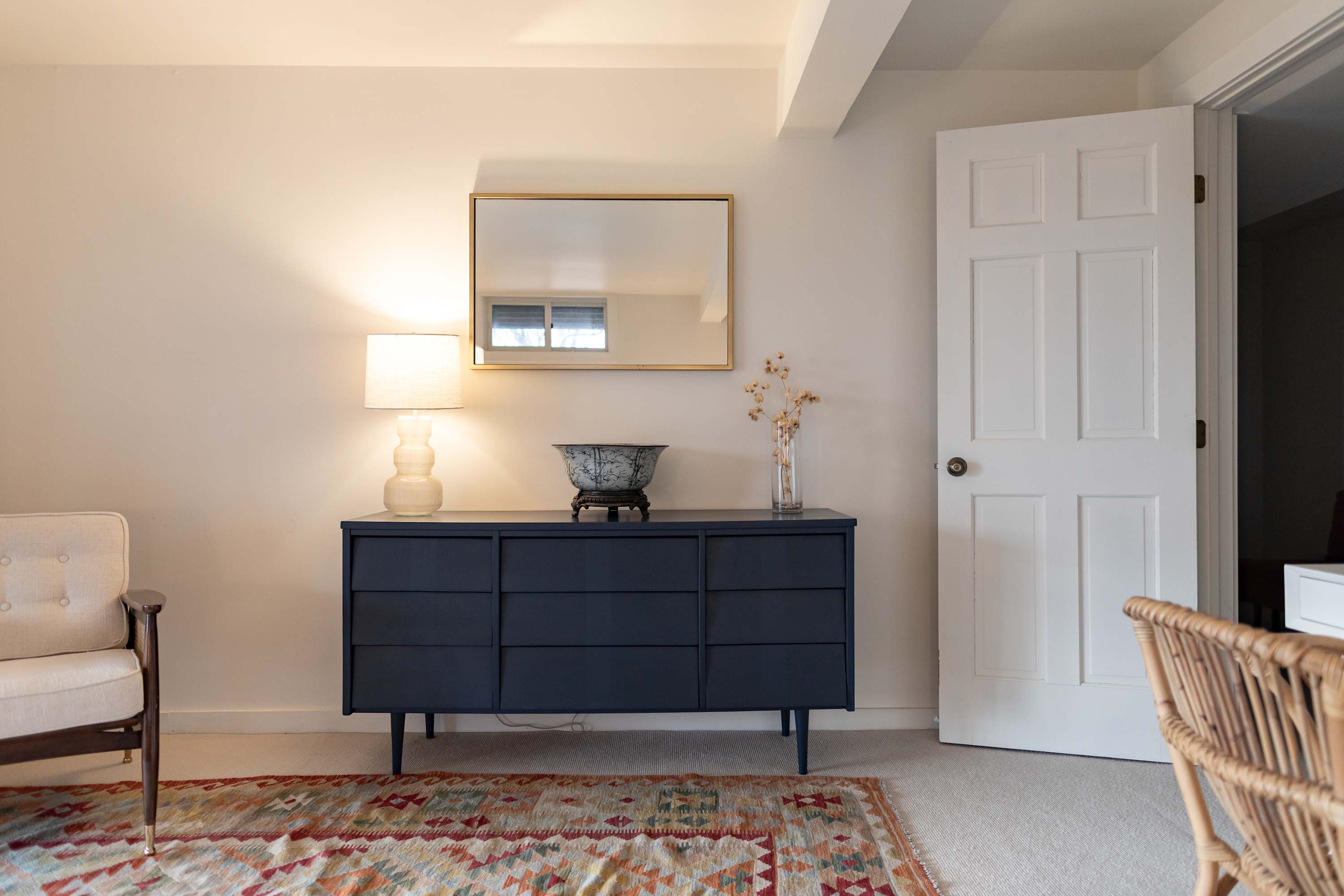 A well-appointed bedroom corner with a dark blue dresser, a white lamp, and a gold-framed mirror above it. A patterned rug covers the light carpet, and a cream armchair and open white door are visible, completing the inviting space.