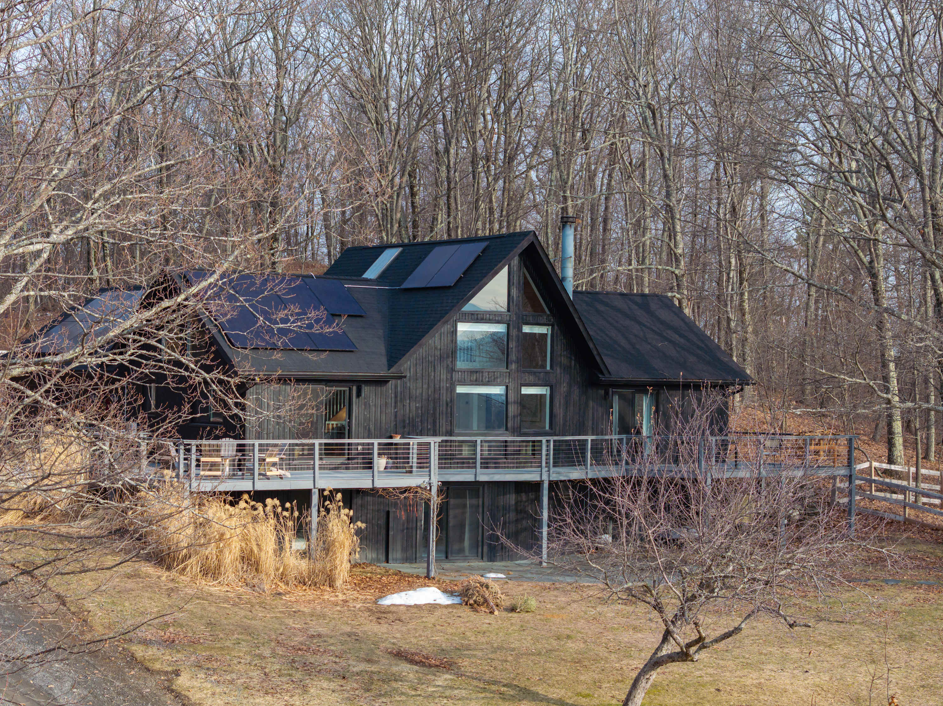A modern black house with large windows and a wrap-around deck is situated on a gentle slope, surrounded by bare trees. Solar panels are visible on the roof, and a gravel path leads up to the property.
