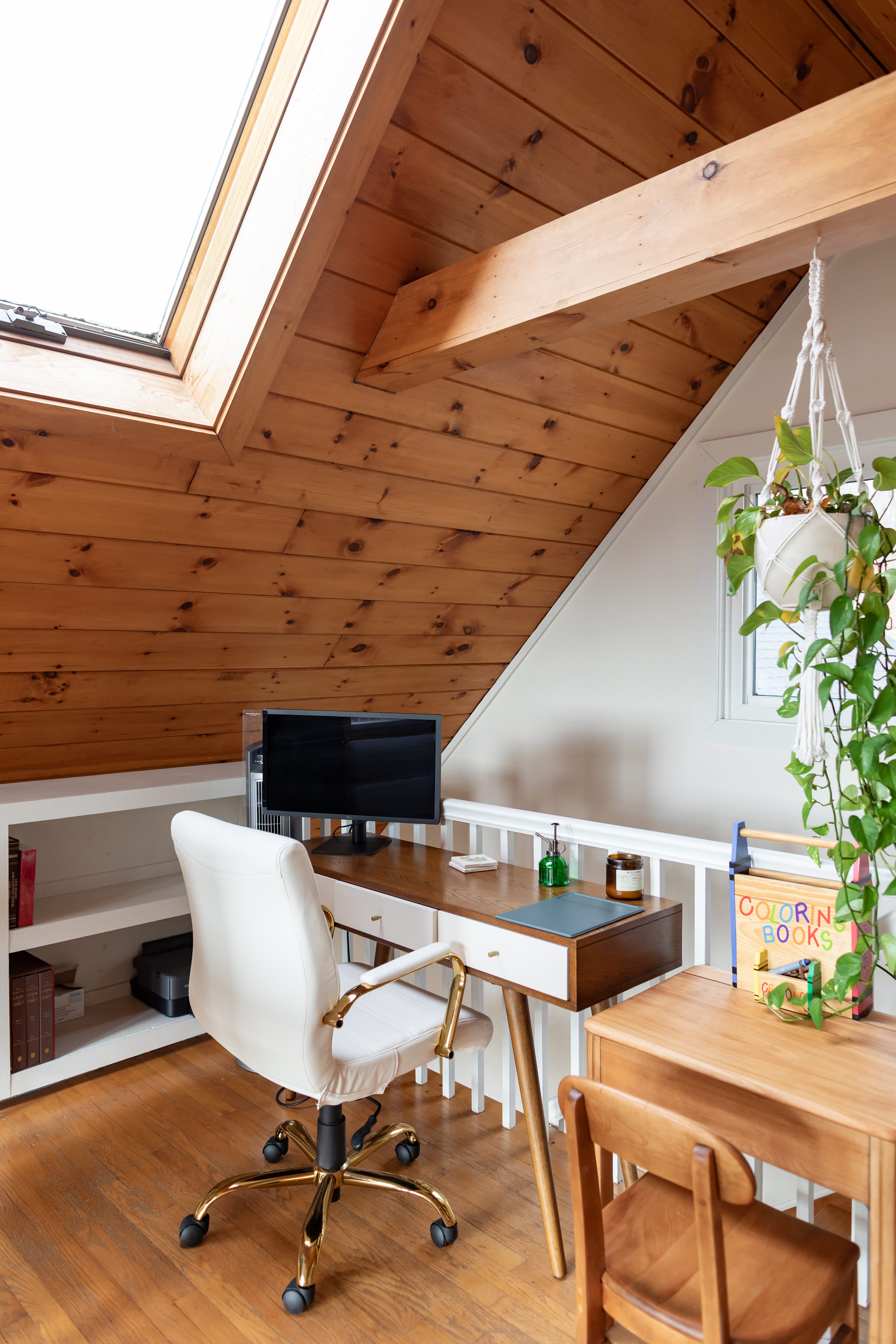 Cozy loft office space with a wooden desk, white and gold office chair, and built-in shelving. A sloped wooden ceiling with a skylight provides natural light, overlooking a children's activity table.