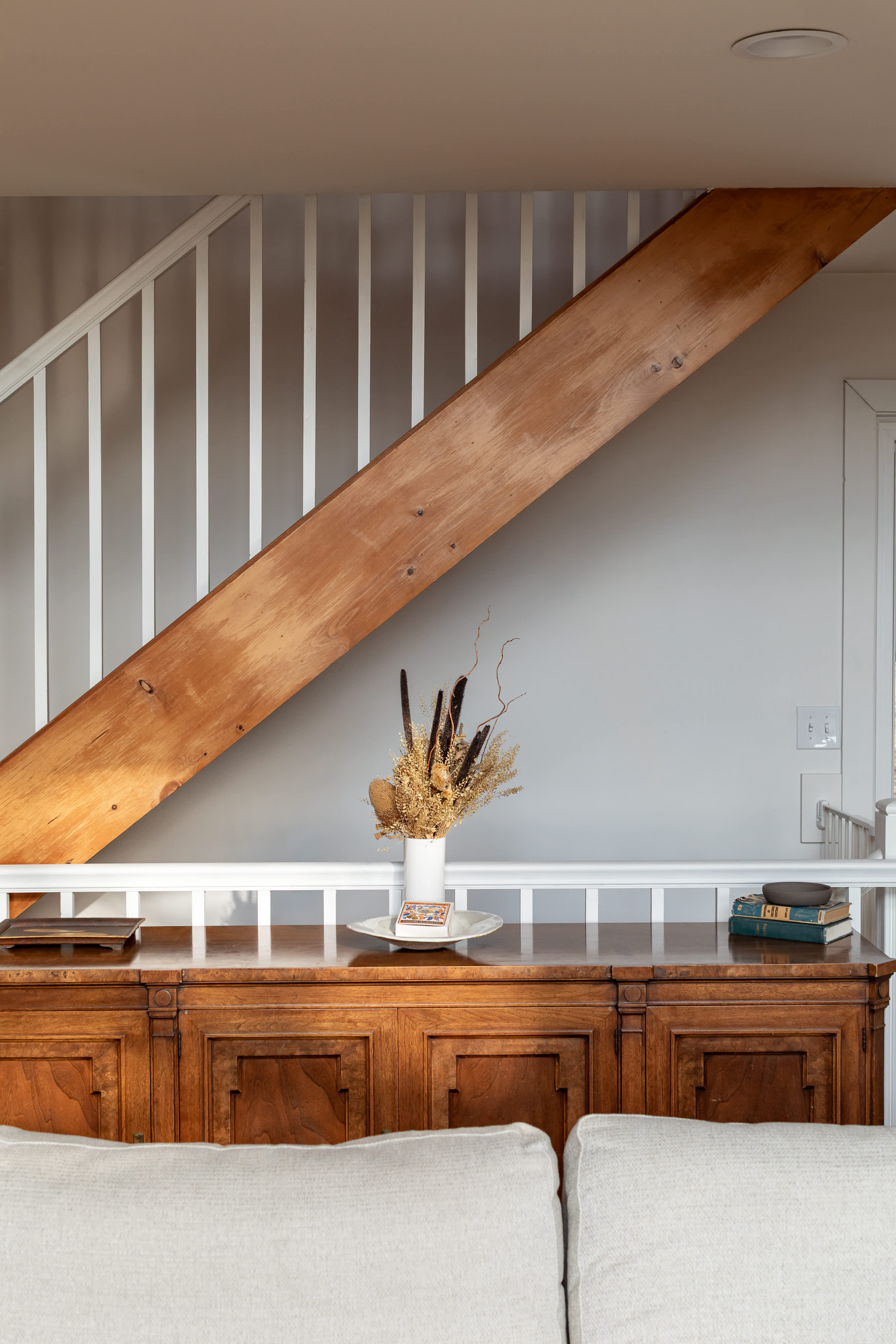 A rustic wooden credenza sits beneath a white-railed staircase, adorned with a dried floral arrangement and vintage books. The wall is painted a light gray, complementing the natural wood tones.