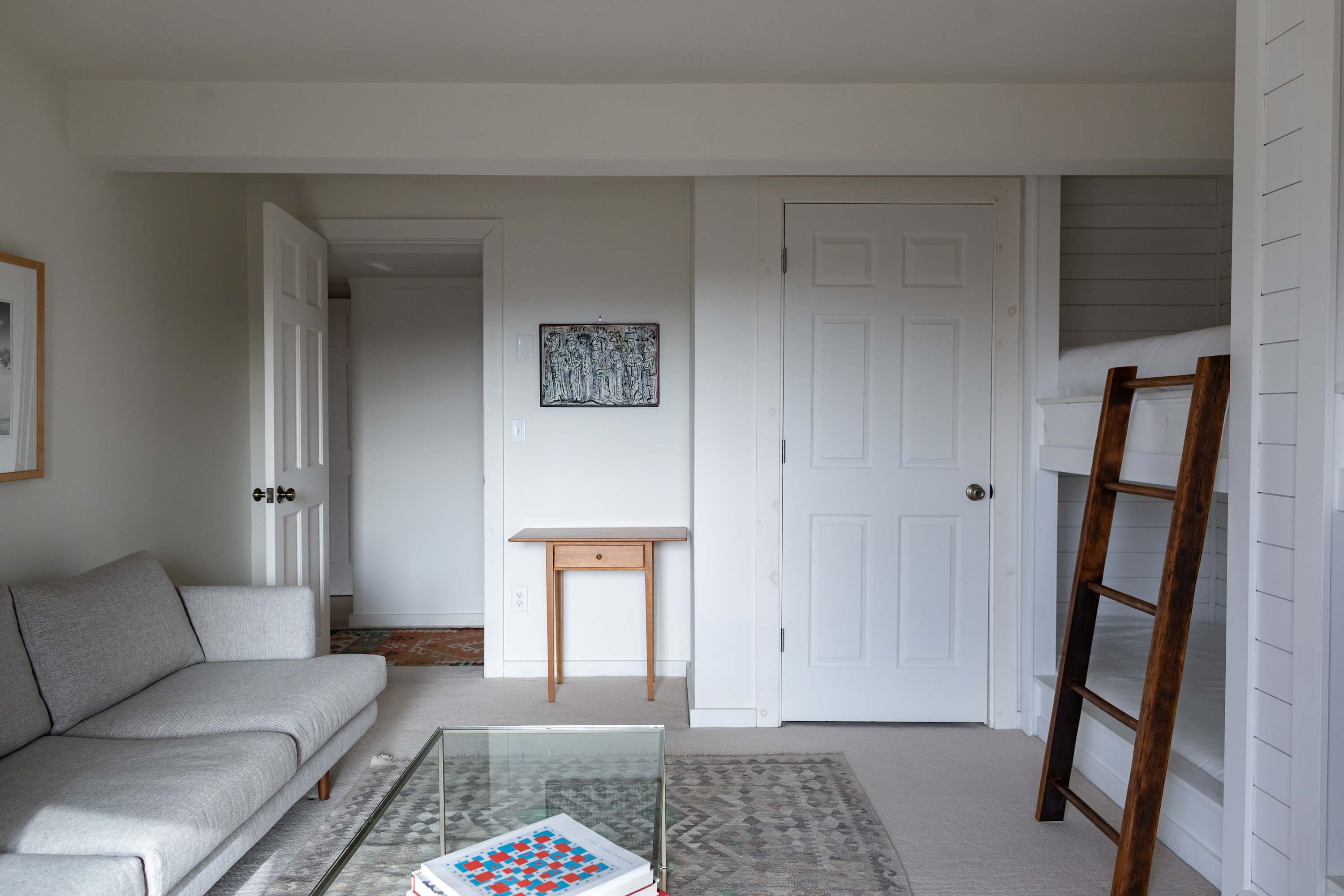 A comfortable living area featuring a light gray sofa, a glass coffee table with books, and a patterned rug on light carpet. Built-in bunk beds are visible on the right, and a white door and open doorway lead to other areas.