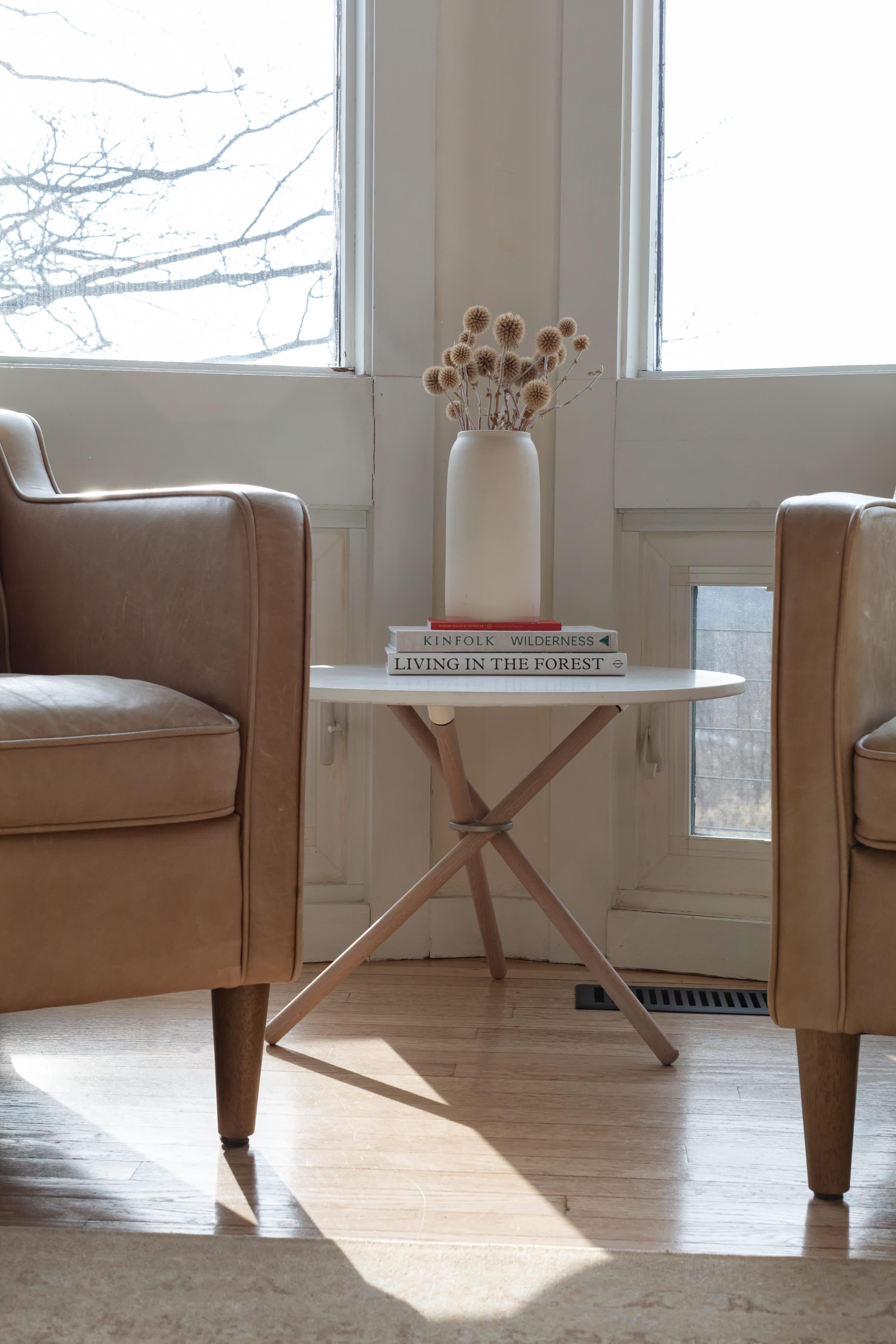 Two comfortable light brown leather armchairs are positioned beside a small round side table, holding books and a vase of dried thistles, all set against a backdrop of large sunlit windows.