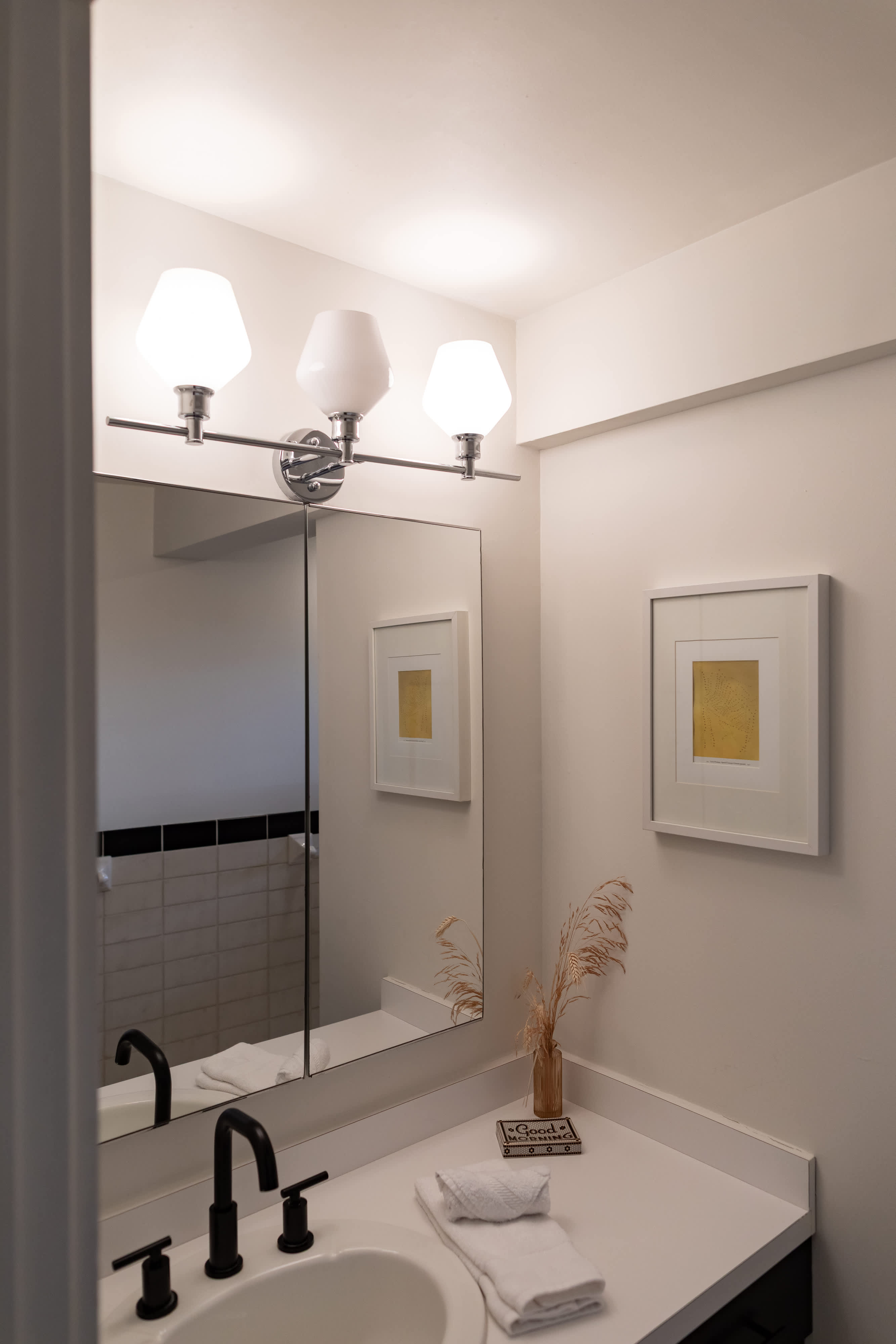 A modern bathroom featuring a white vanity with a sleek black faucet and white sink, a large mirror, and a three-light vanity fixture. White subway tiles are visible on the lower wall, adding a clean, classic touch.