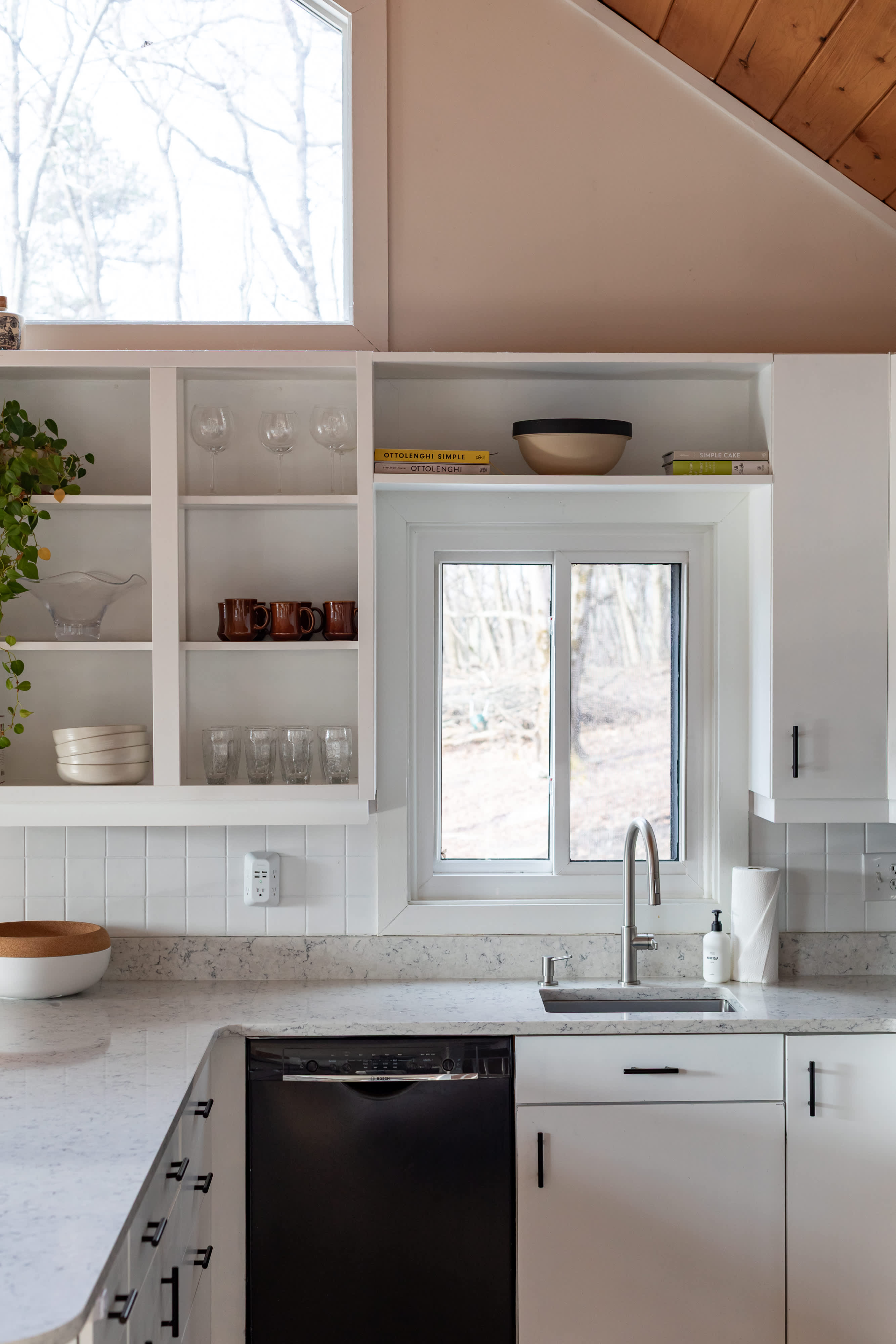 A bright kitchen features white shaker-style cabinets, open shelving displaying glassware and bowls, a white subway tile backsplash, and a light-colored quartz countertop with a stainless steel sink under a window.