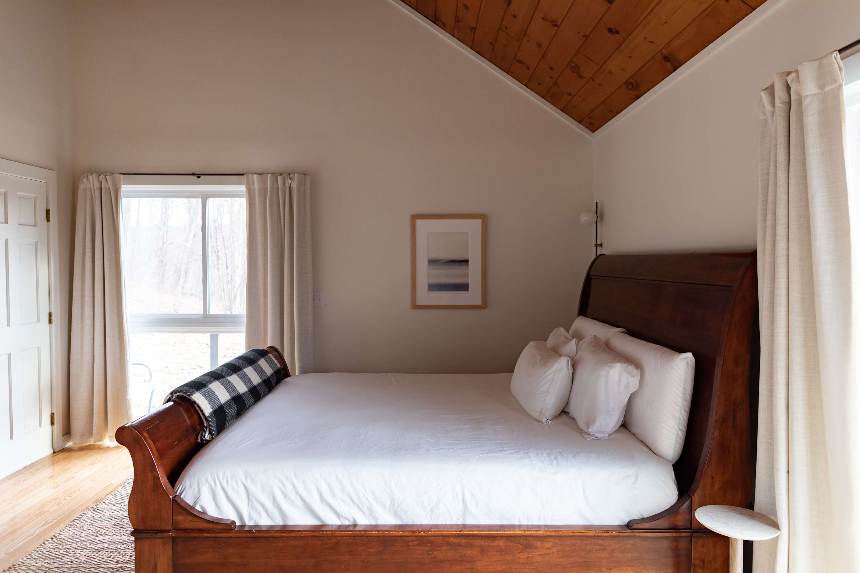 Serene bedroom featuring a dark wooden sleigh bed with crisp white bedding and a plaid throw blanket. The room boasts a vaulted wooden ceiling and a large window with light-colored curtains.