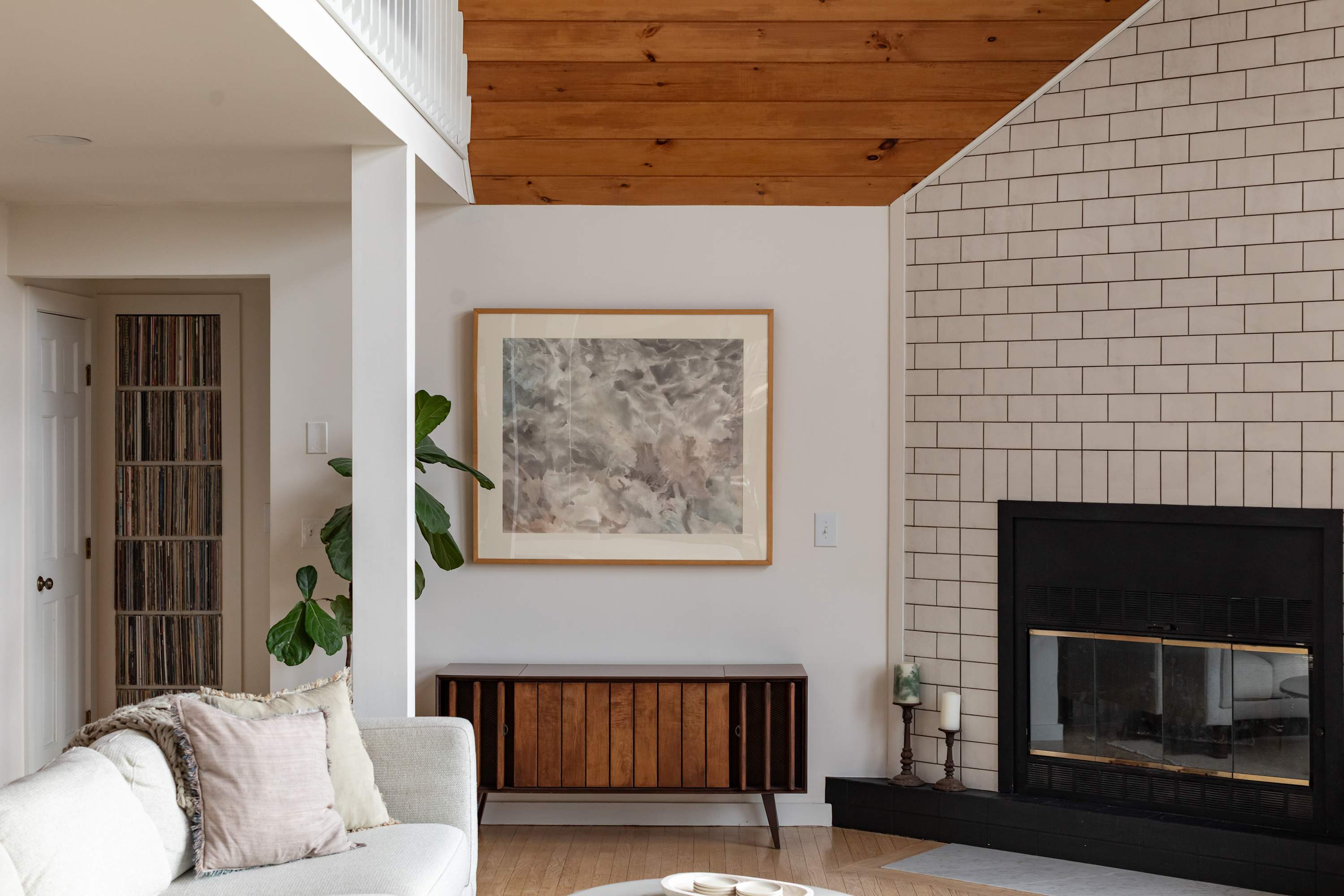 A cozy living room features a white sofa with throw pillows, a mid-century modern credenza under abstract art, and a fireplace with white subway tile surround. A wooden beam ceiling is visible above.