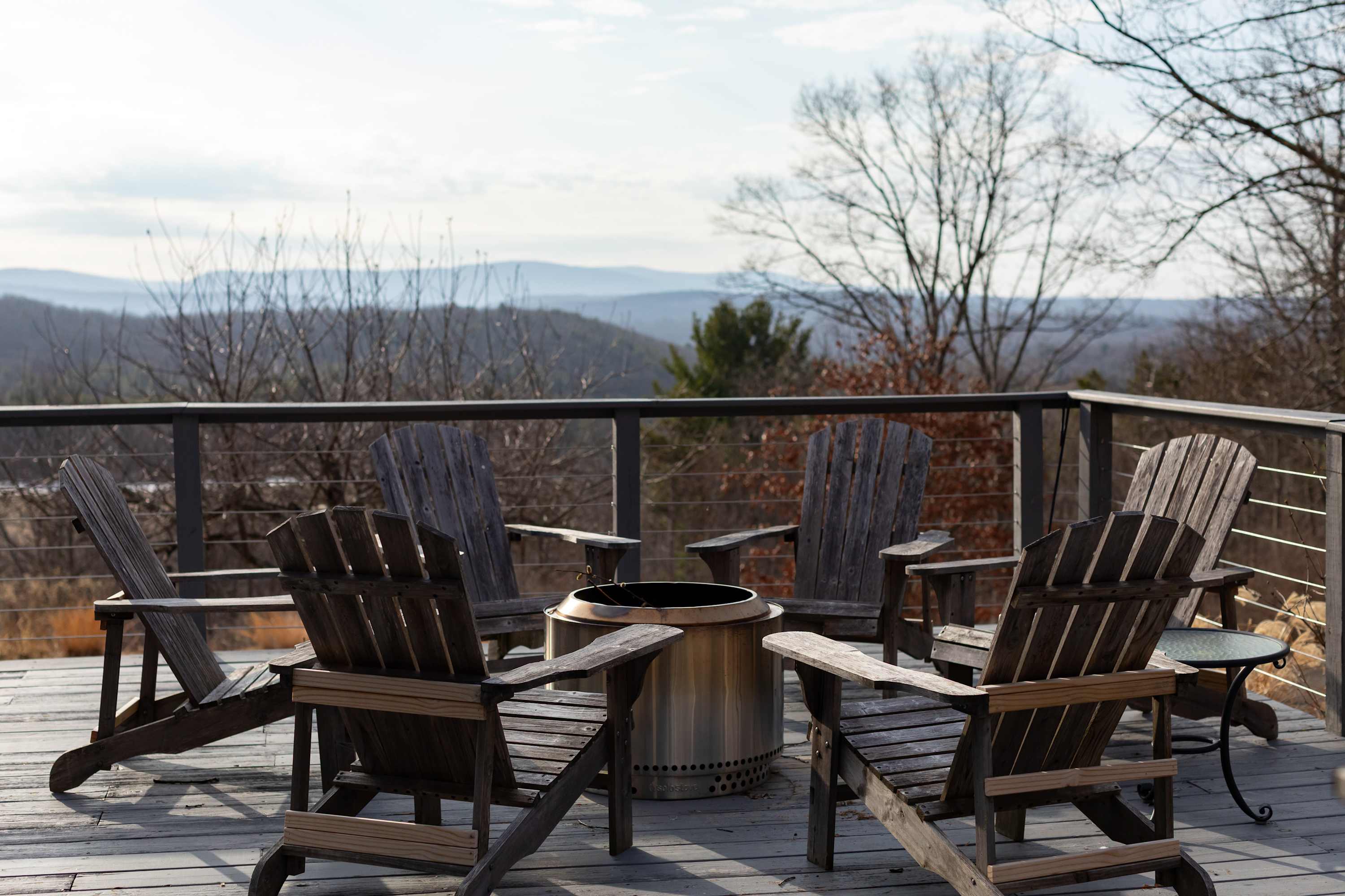 A wooden deck features a circular fire pit surrounded by several rustic wooden Adirondack chairs, offering panoramic views of distant mountains and a hazy sky.