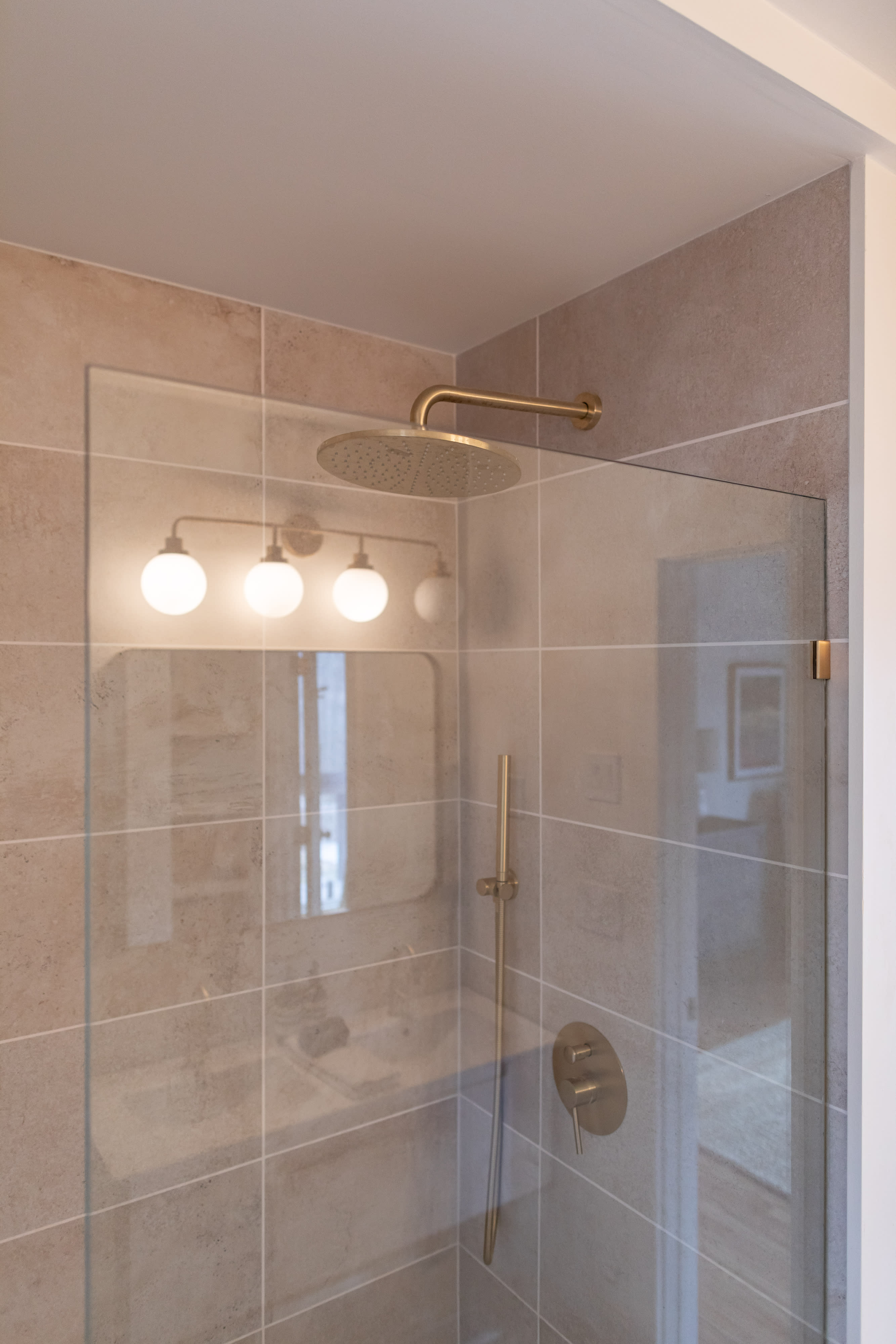 Contemporary shower with a clear glass enclosure, gold rain shower head, and matching gold controls. The walls are tiled in a light, neutral tone, reflecting the vanity lights from outside the shower.