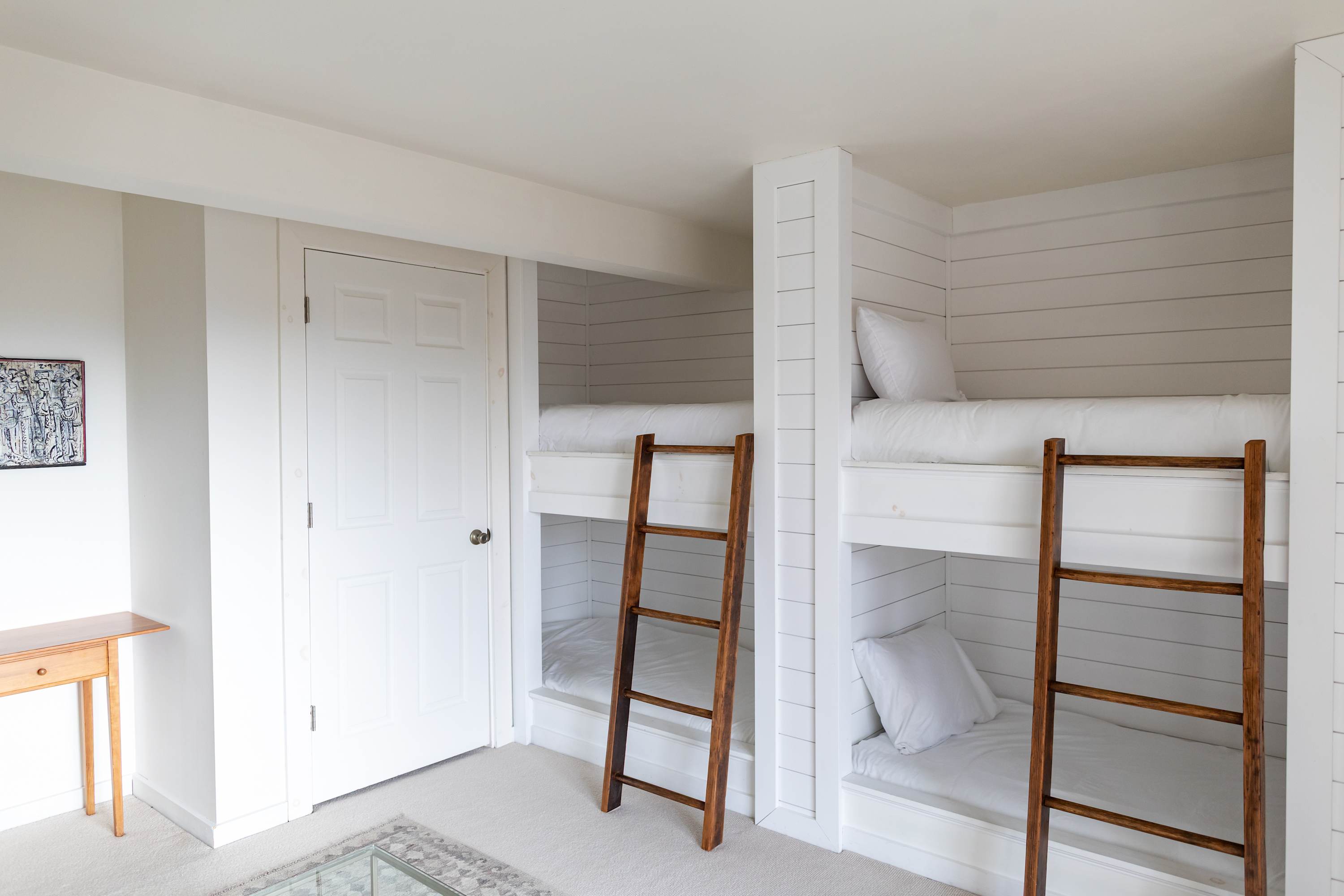 A bright bunk room with two sets of built-in white shiplap bunk beds, each featuring white bedding and a sturdy wooden ladder. A white paneled door is visible on the left, and the room is carpeted in a light neutral tone.