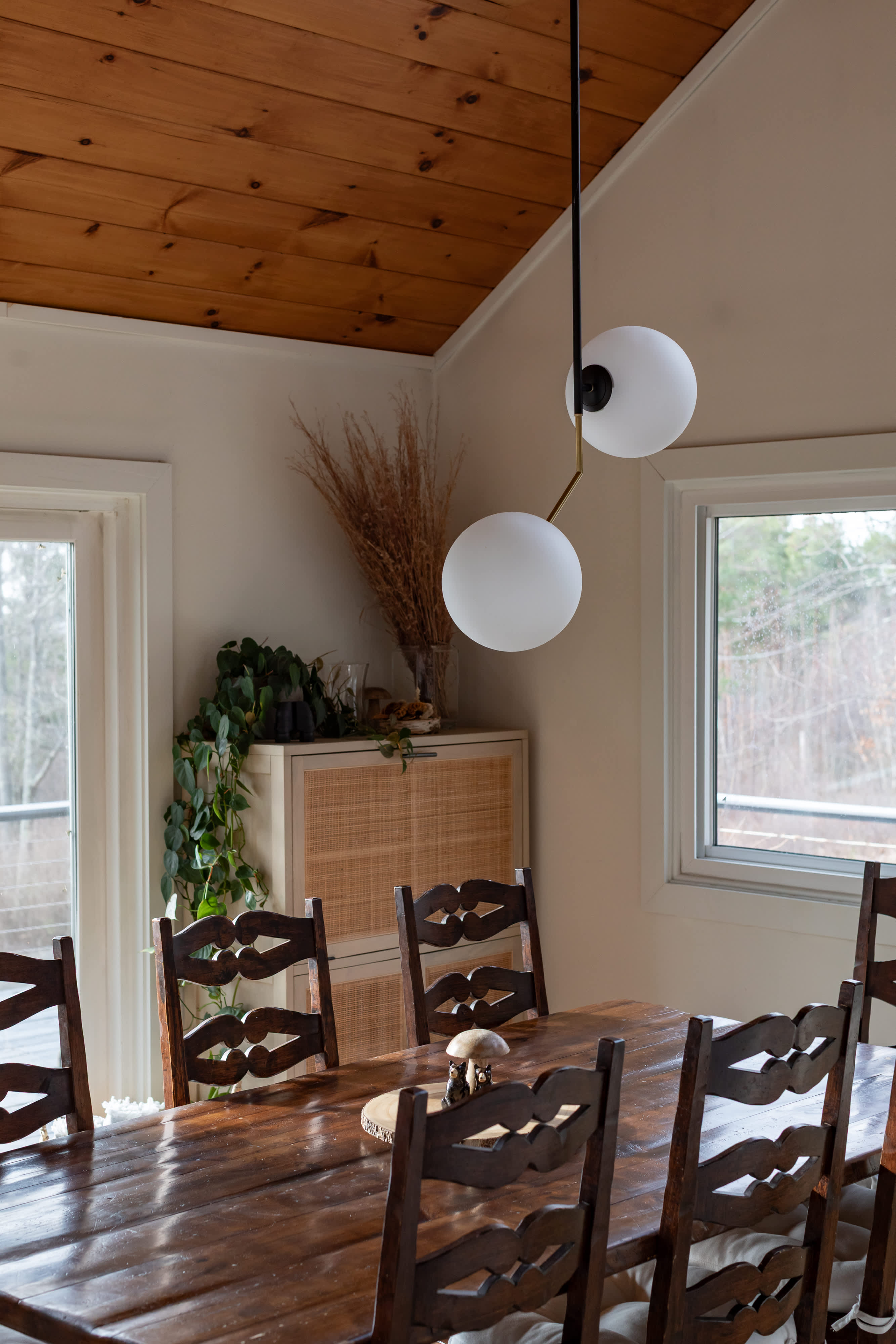 A rustic dining area features a large, dark wooden table surrounded by matching wooden chairs, illuminated by a modern pendant light with two spherical globes, with a rattan-front cabinet in the background.