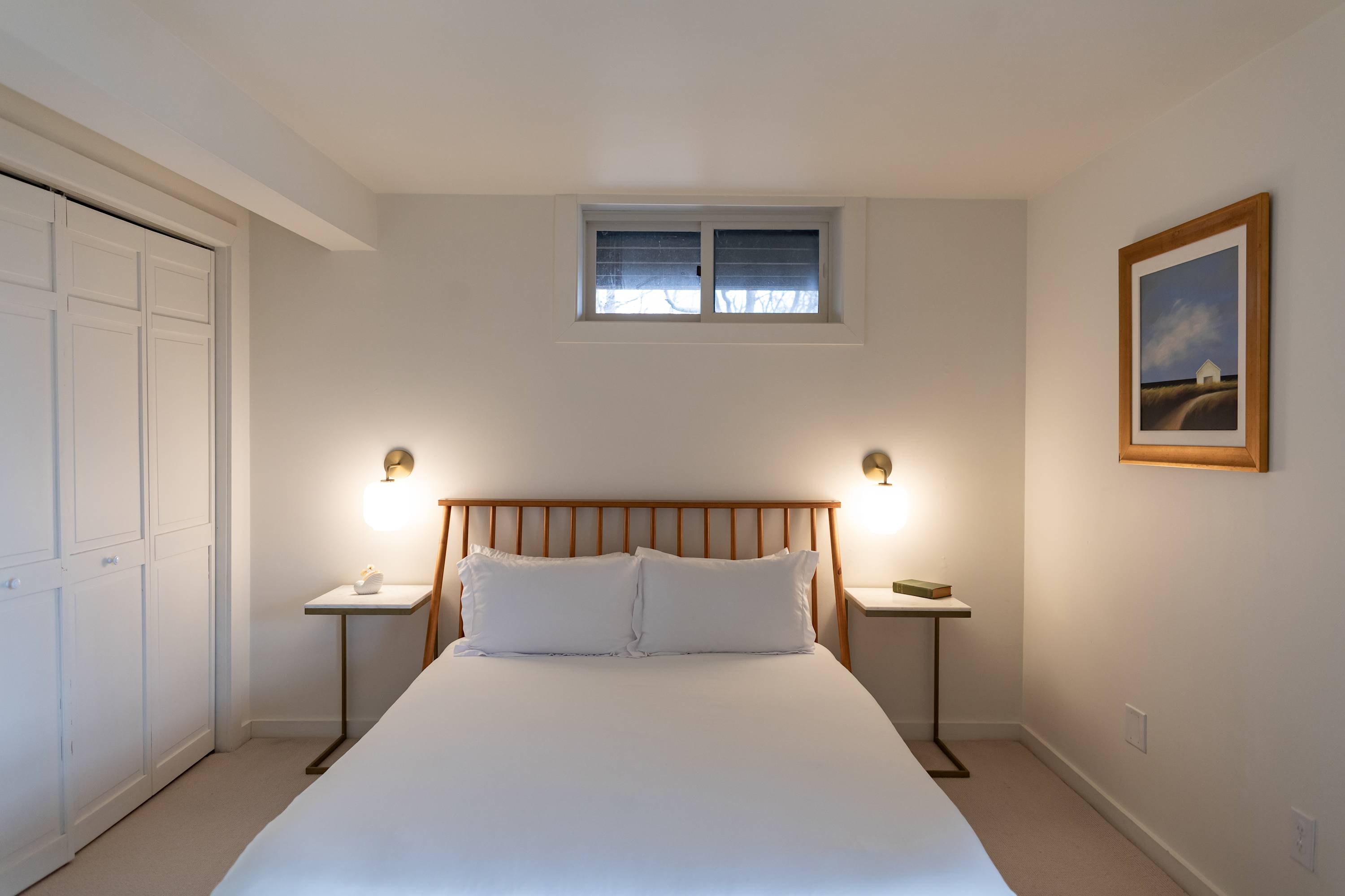 A serene bedroom with a wooden bed frame, slatted headboard, and white bedding, flanked by two modern side tables with brass wall sconces. A small window is above the bed, and a white bi-fold closet is on the left, providing storage.