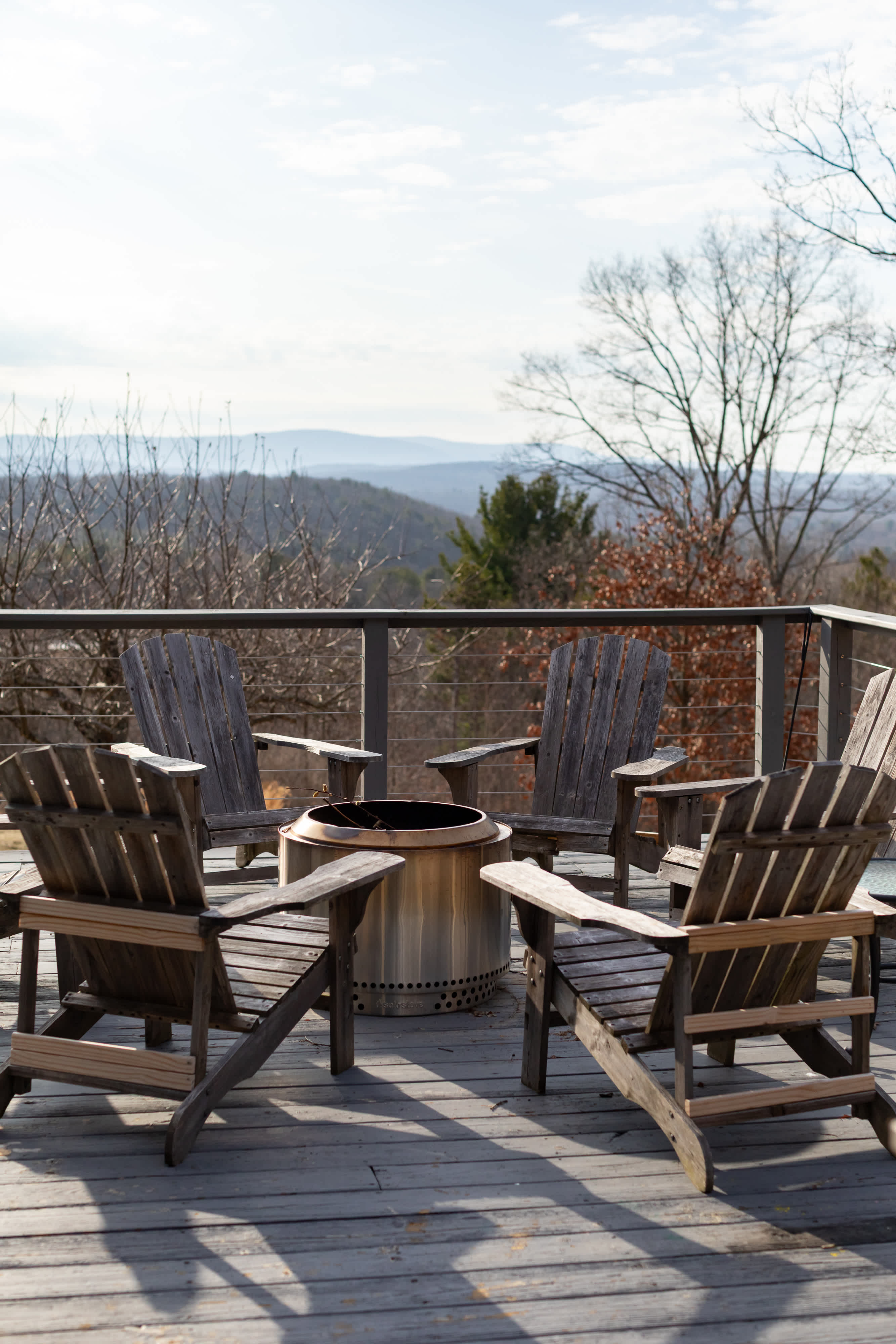 A wooden deck features four rustic Adirondack chairs arranged around a modern, stainless steel fire pit, offering expansive mountain views in the distance.