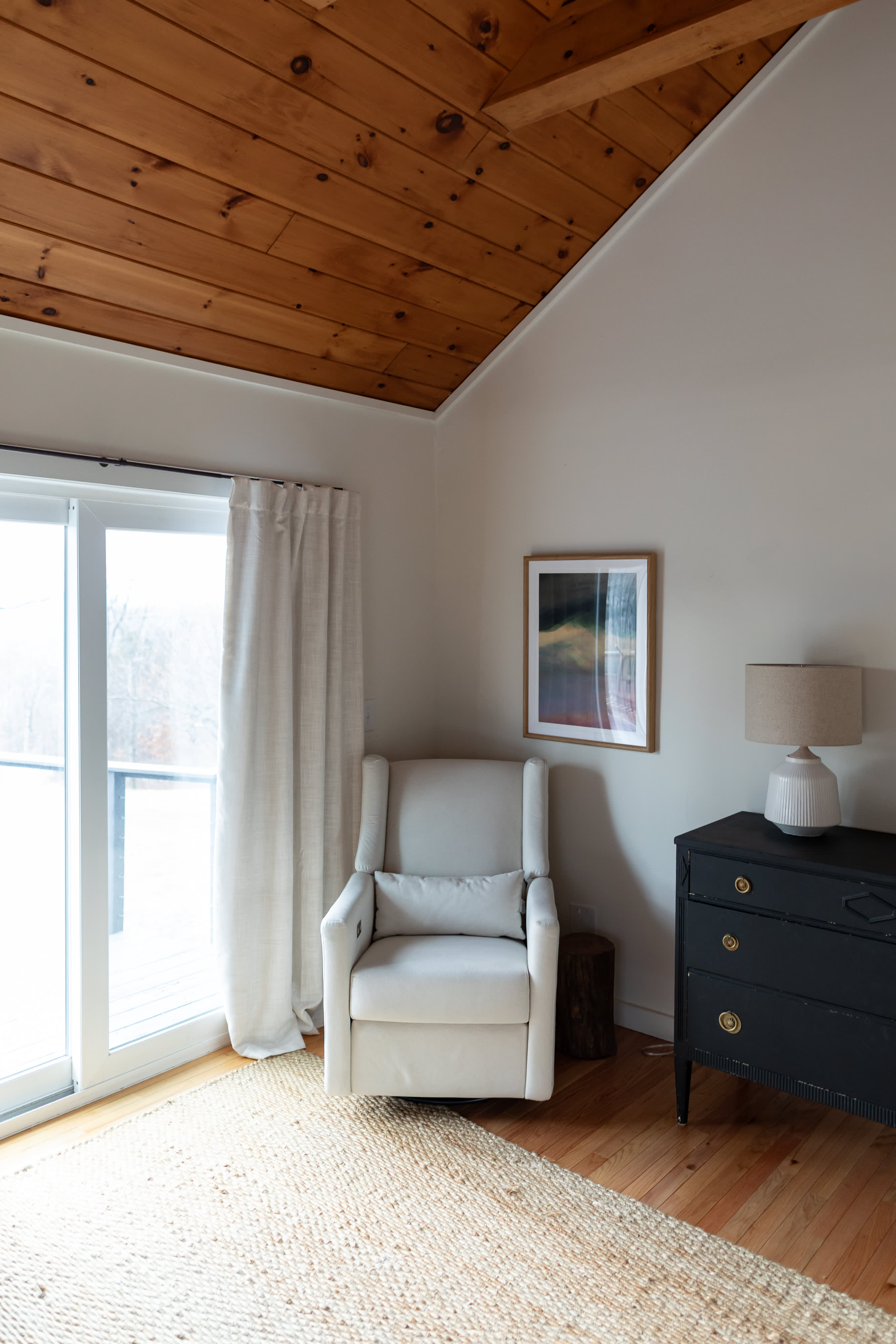 Bright bedroom corner with a comfortable white recliner, a black dresser with a lamp, and a large sliding glass door leading outdoors. The room features a vaulted wooden ceiling and a woven rug over hardwood floors.