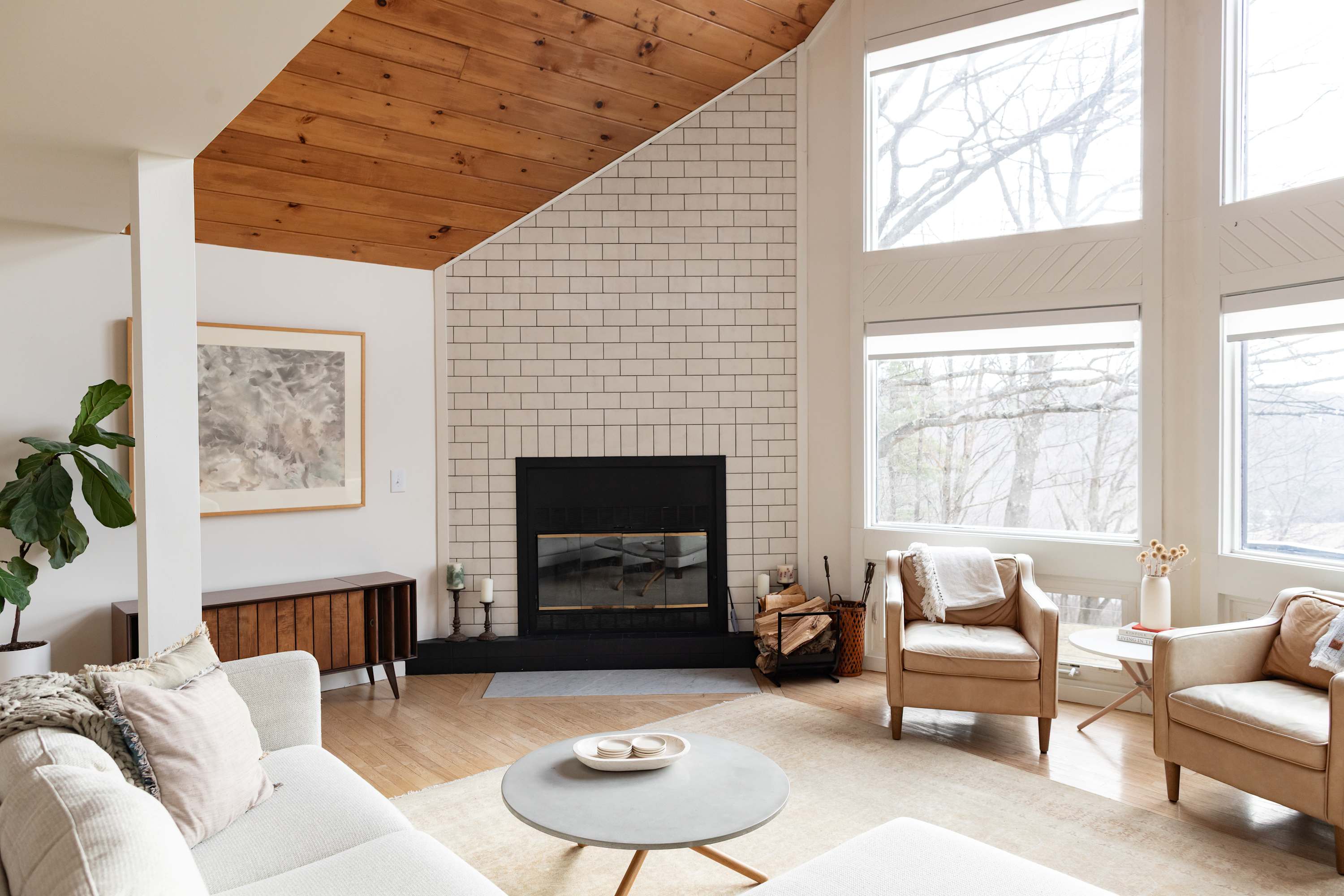 A spacious living room boasts a vaulted wooden ceiling and a fireplace with a white subway tile surround, furnished with a light-colored sectional sofa and two armchairs, all bathed in natural light from large windows.