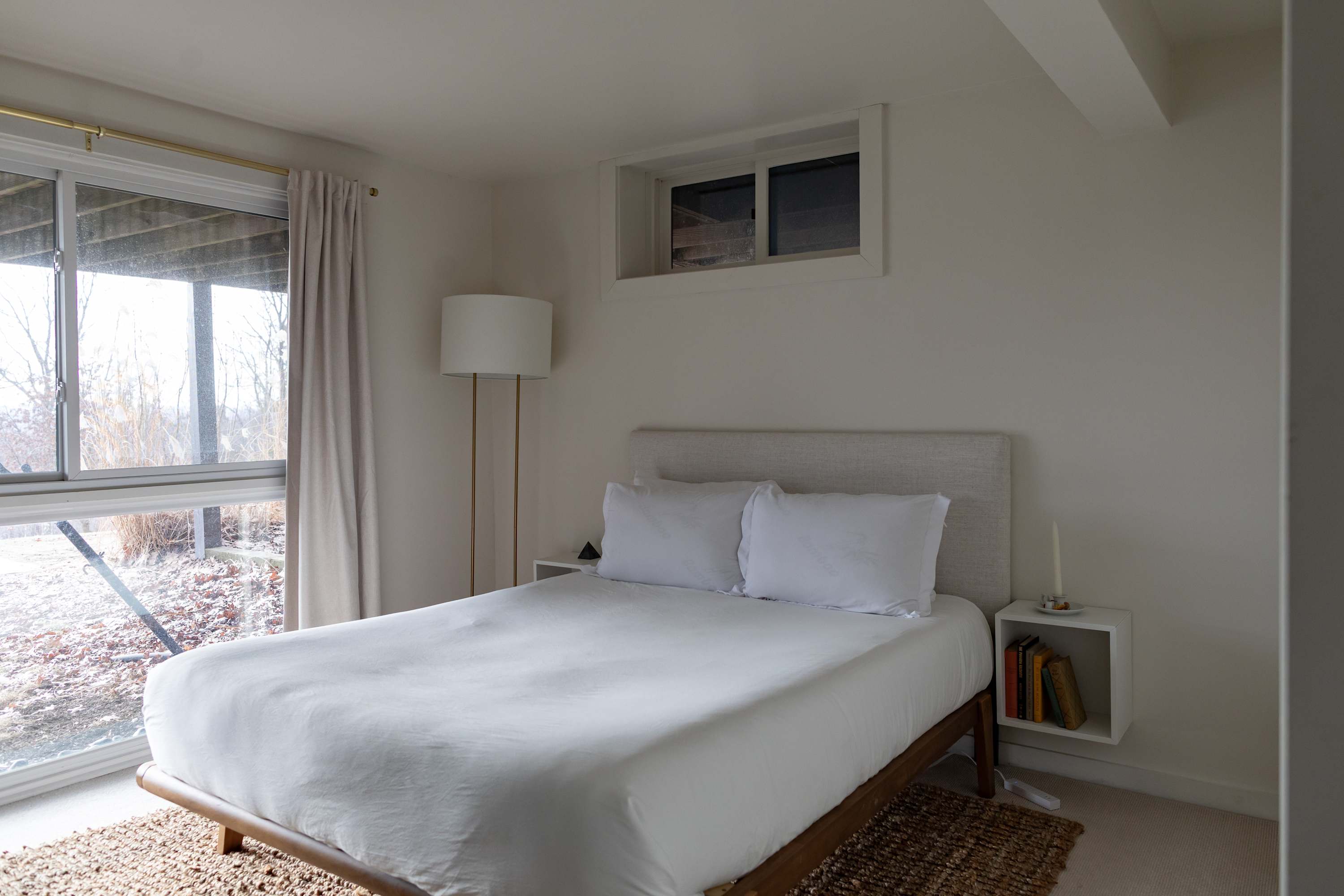 Inviting bedroom with a bed featuring a light upholstered headboard and crisp white bedding. A large window with light curtains offers views, complemented by a floor lamp and a small bedside shelf with books.