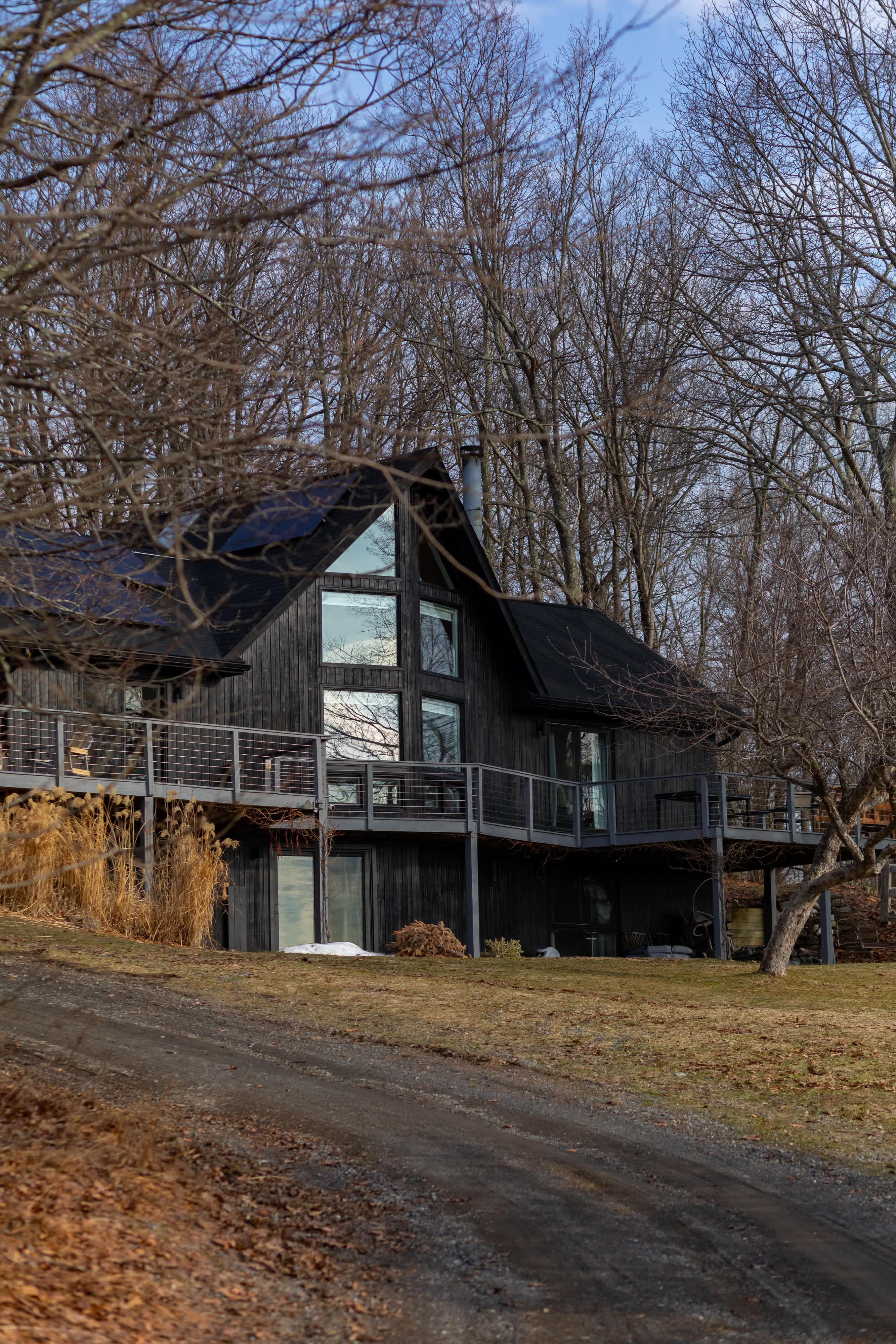 Modern black-sided house with large windows and a wrap-around deck, nestled among bare trees on a sloped, grassy terrain with a dirt driveway. Solar panels are visible on the roof, adding to its contemporary design.