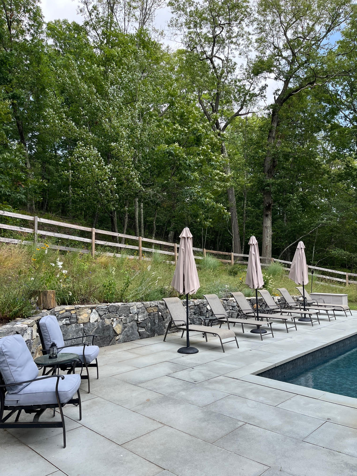 Expansive stone patio adjacent to an inground swimming pool, furnished with comfortable grey cushioned chairs, a small side table, and several lounge chairs with beige umbrellas. A stone retaining wall and a wooden fence separate the leisure area from a lush, green forest.