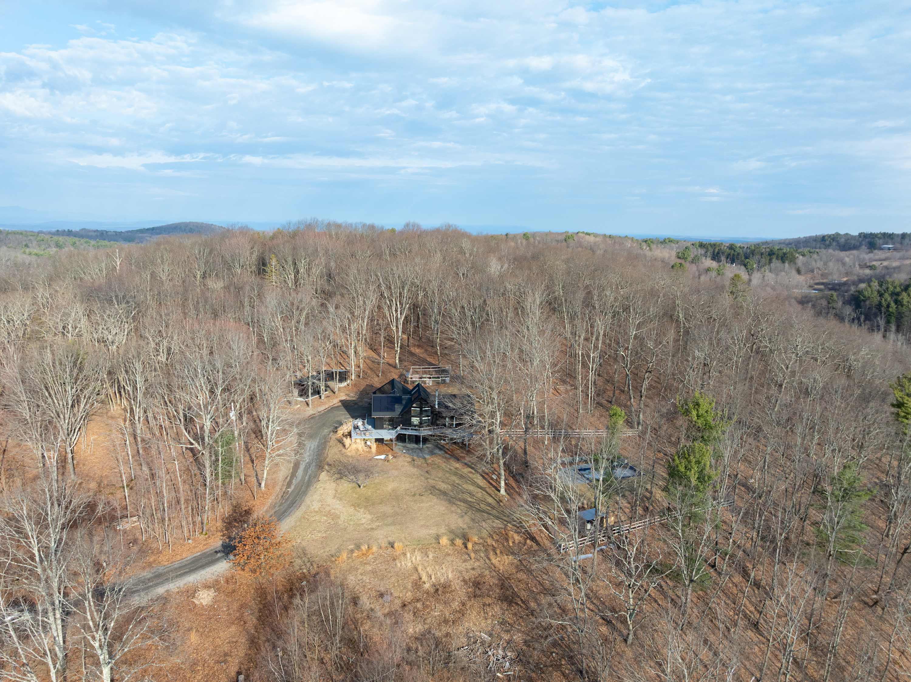 Sweeping aerial view of a modern black house with solar panels, nestled within a vast, bare forest on a gently sloped landscape, with a winding dirt road leading to the property. Distant mountains are visible under a partly cloudy sky, showcasing the secluded setting.