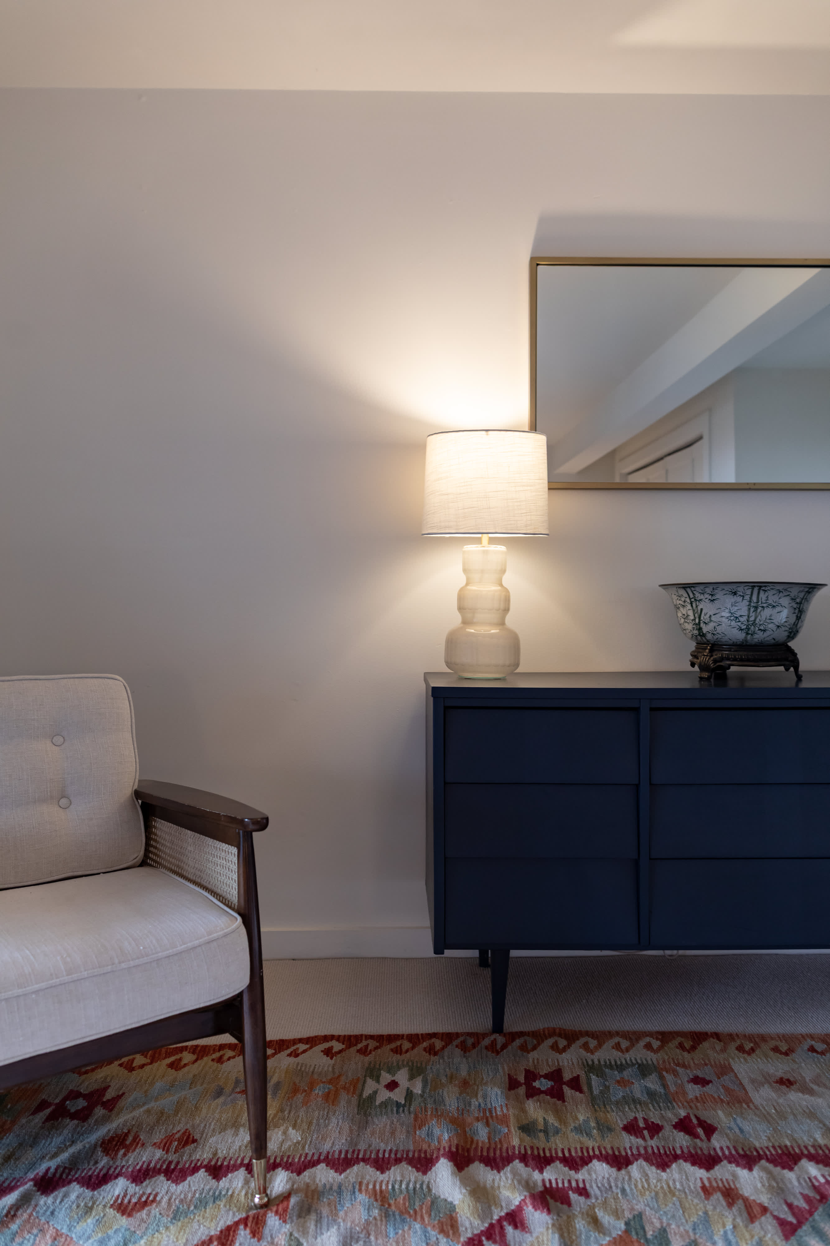 A stylish corner of a bedroom featuring a dark blue dresser with a white lamp, a gold-framed mirror, and a patterned rug on light carpet. A cream-colored armchair with wooden accents is visible on the left, offering a comfortable seating area.