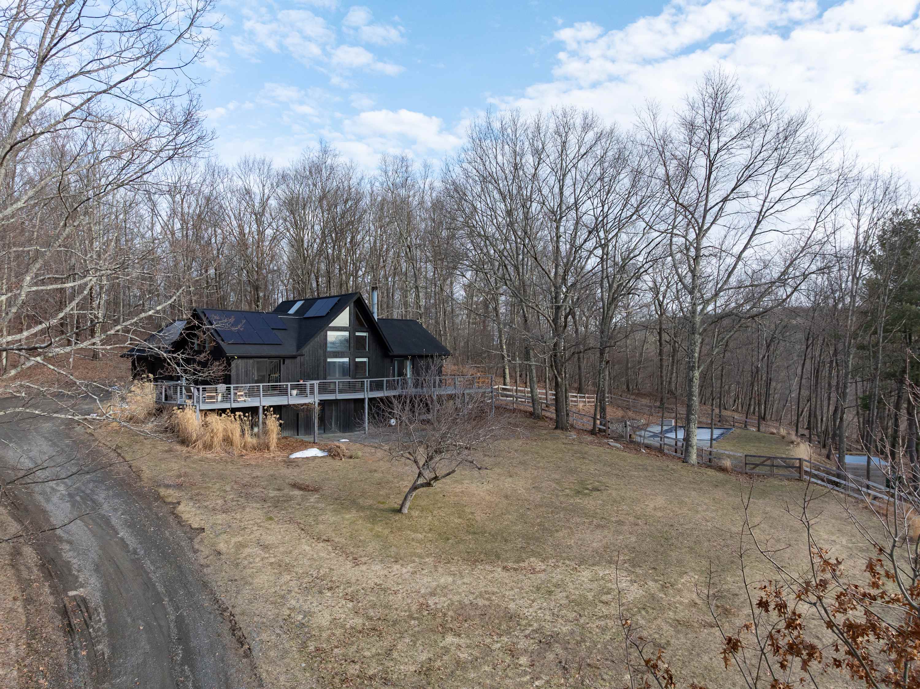 Wide ground-level shot of the modern black house featuring a wrap-around deck and prominent large windows, surrounded by bare trees and a sloped, grassy yard. A dirt driveway leads up to the property, with a fenced pool area visible in the distance.
