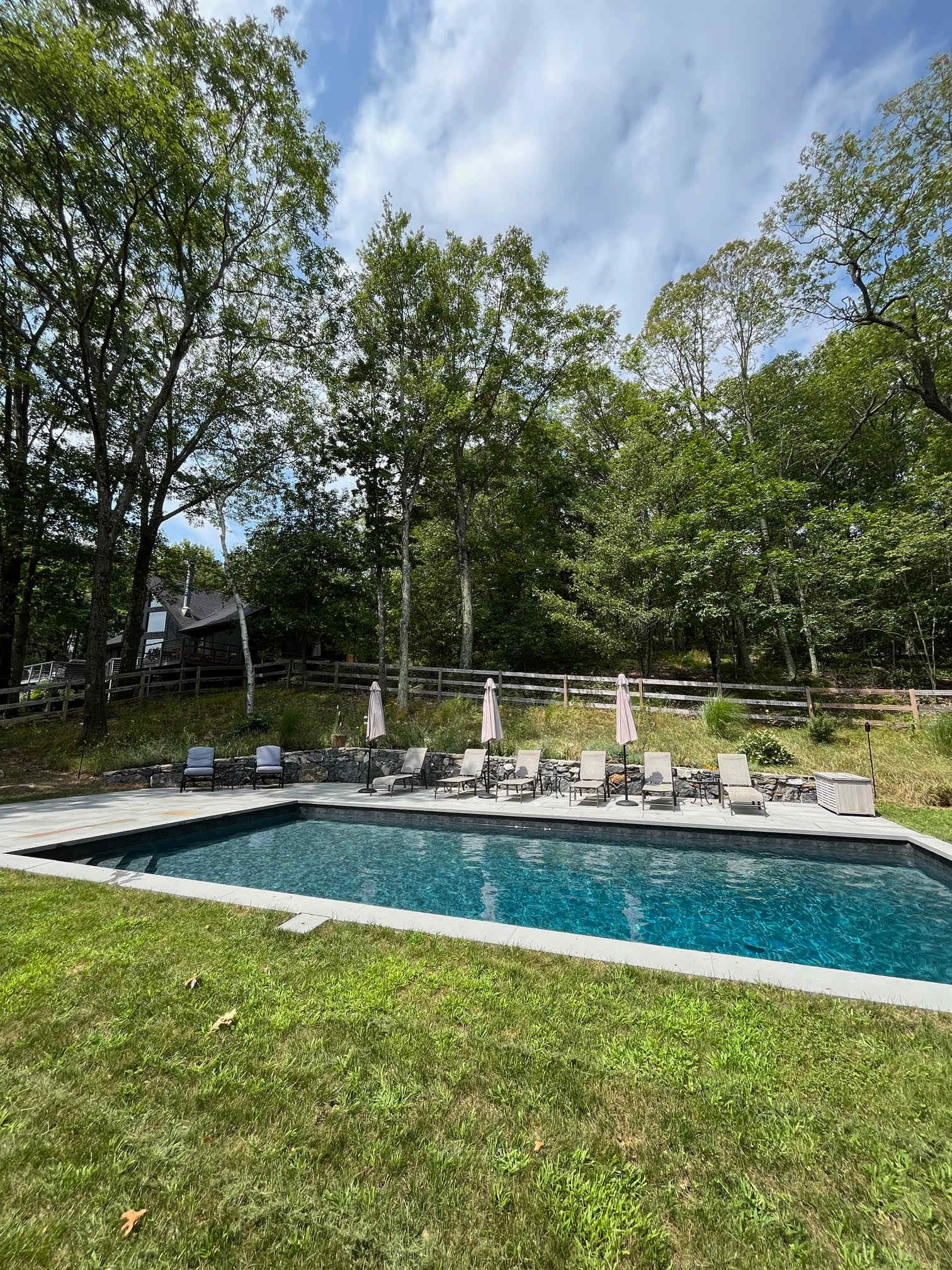 Rectangular inground swimming pool with dark blue water, surrounded by a light-colored stone patio and vibrant green grass, featuring multiple lounge chairs and beige umbrellas. A dense forest and a rustic wooden fence provide a natural backdrop.
