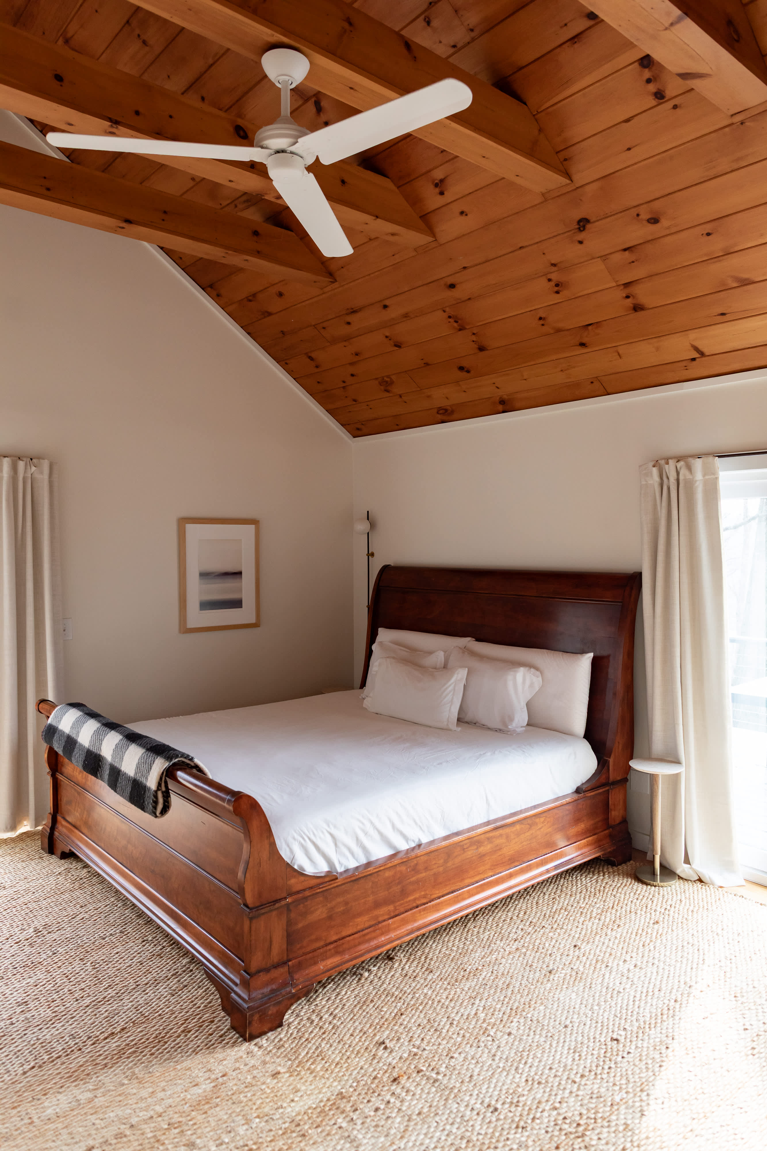 Spacious bedroom with a prominent dark wooden sleigh bed, white bedding, and a plaid blanket. A vaulted wooden ceiling with a ceiling fan adds to the rustic charm, complemented by a woven rug and large windows.