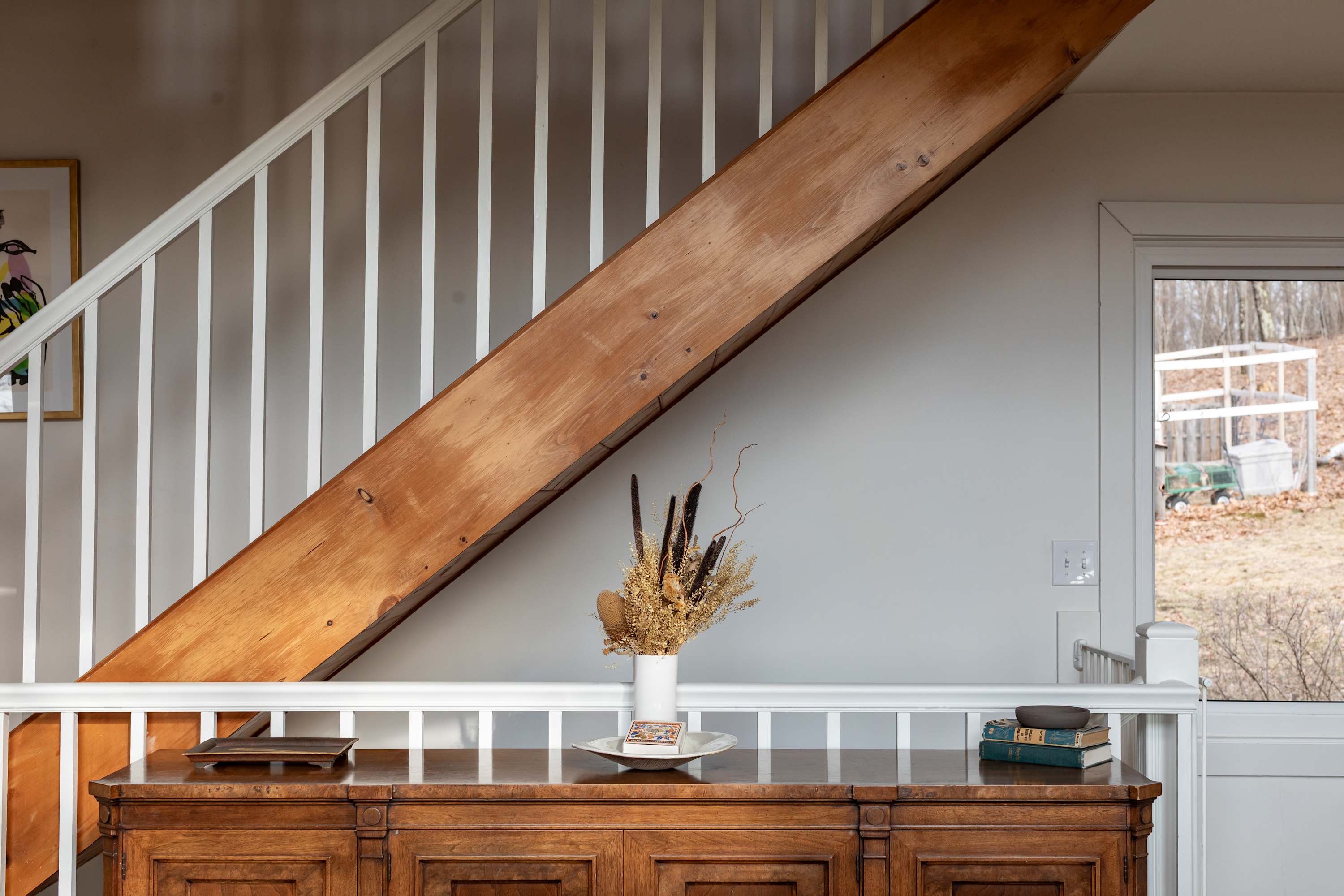 A striking wooden staircase with white balusters and a large exposed beam ascends above a rustic wooden console table, adorned with a vase of dried botanicals and stacked books.