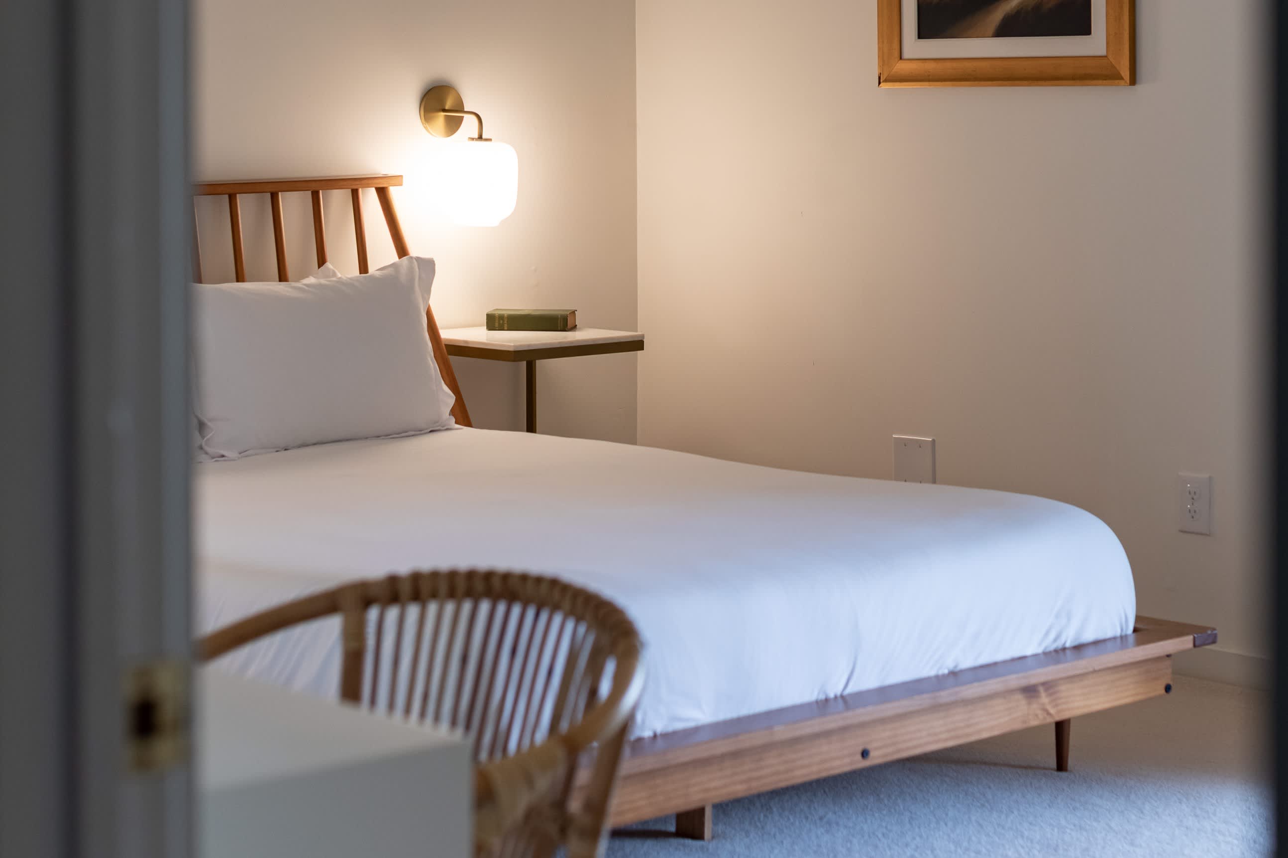 A cozy bedroom viewed through a doorway, featuring a wooden bed frame with a slatted headboard and crisp white bedding. A small side table with a brass wall sconce and a book is positioned next to the bed, offering warm lighting.