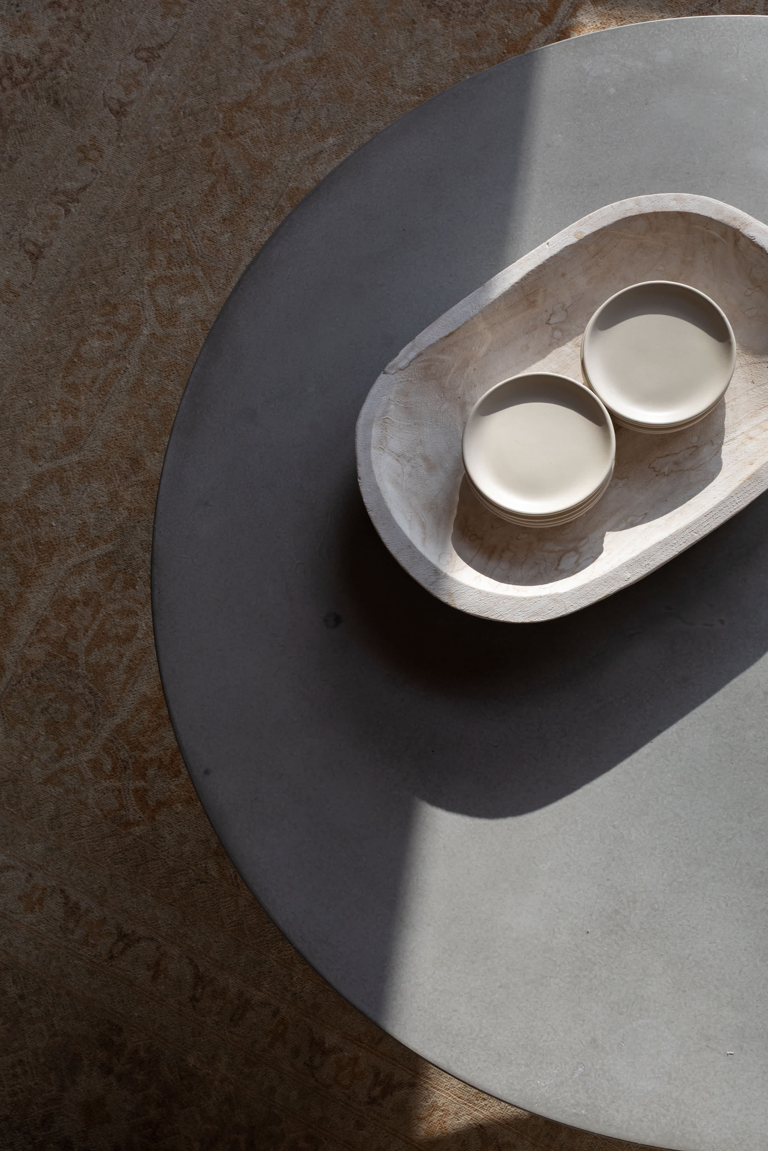 Overhead view of a round, light gray coffee table holding a wooden bowl with two small, light-colored plates. Sunlight casts a shadow across the table and a patterned rug.