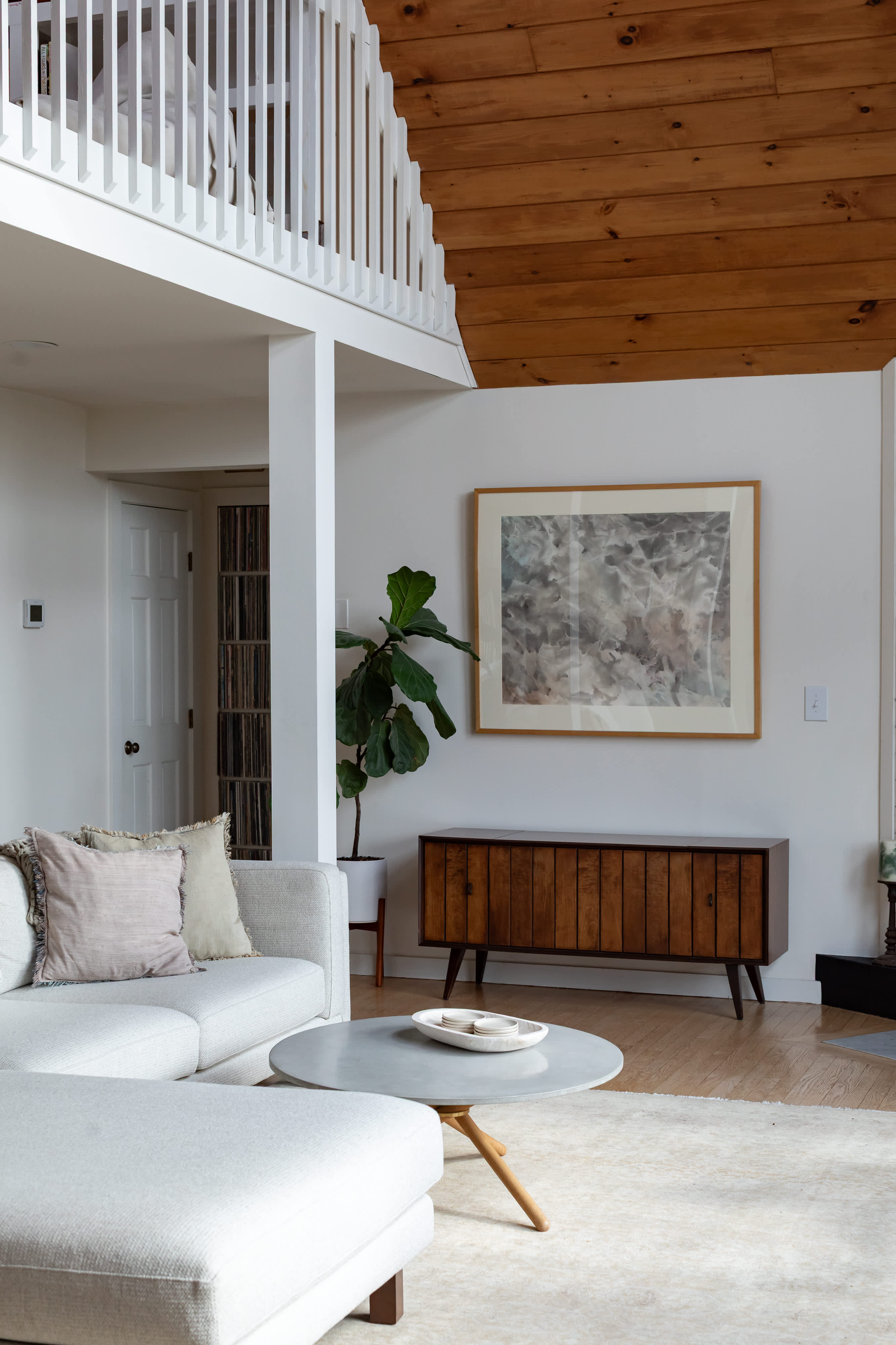 Bright living room featuring a white sectional sofa, a round concrete coffee table, and a wooden credenza under abstract art. A loft with white railings overlooks the space, which has a warm wooden ceiling.