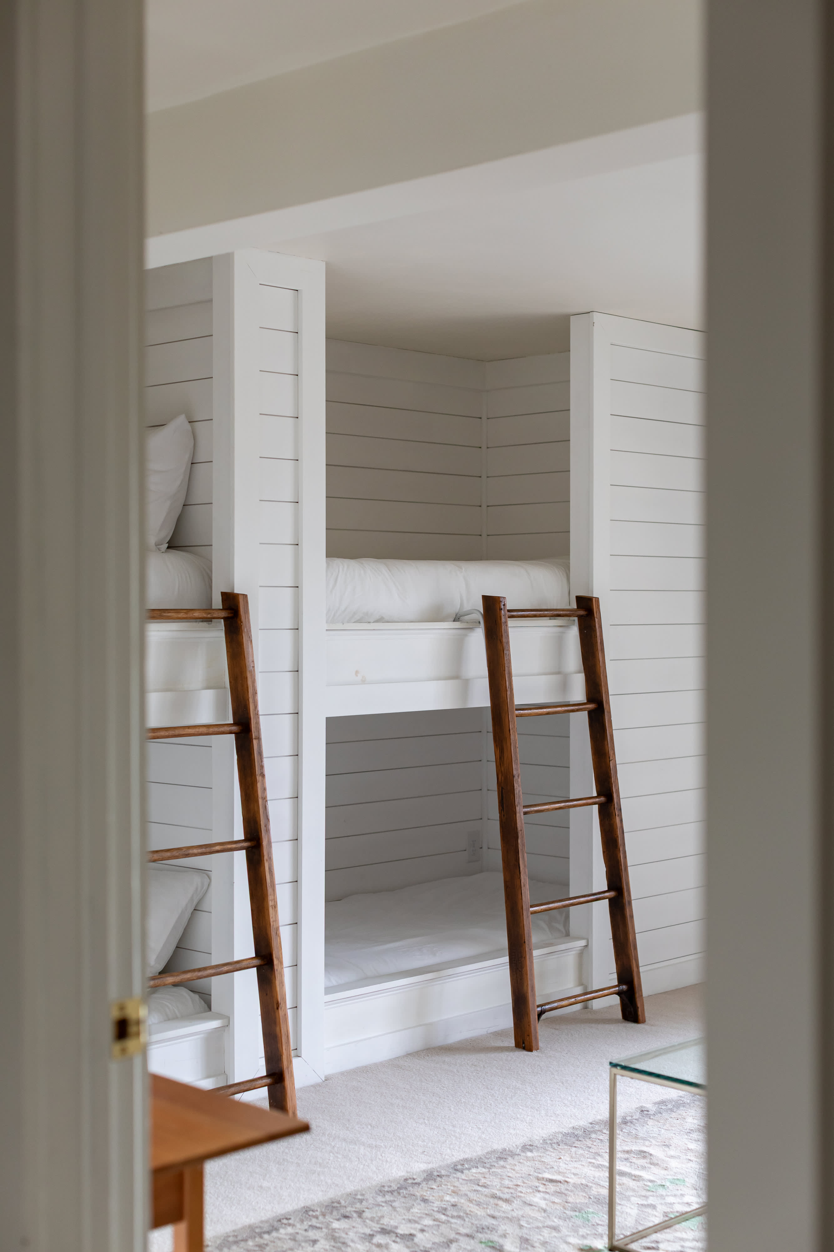 A cozy bunk room featuring built-in white shiplap bunk beds with crisp white bedding, accessed by rustic wooden ladders. The room has light-colored carpet and is seen through a doorway, creating a private sleeping nook.