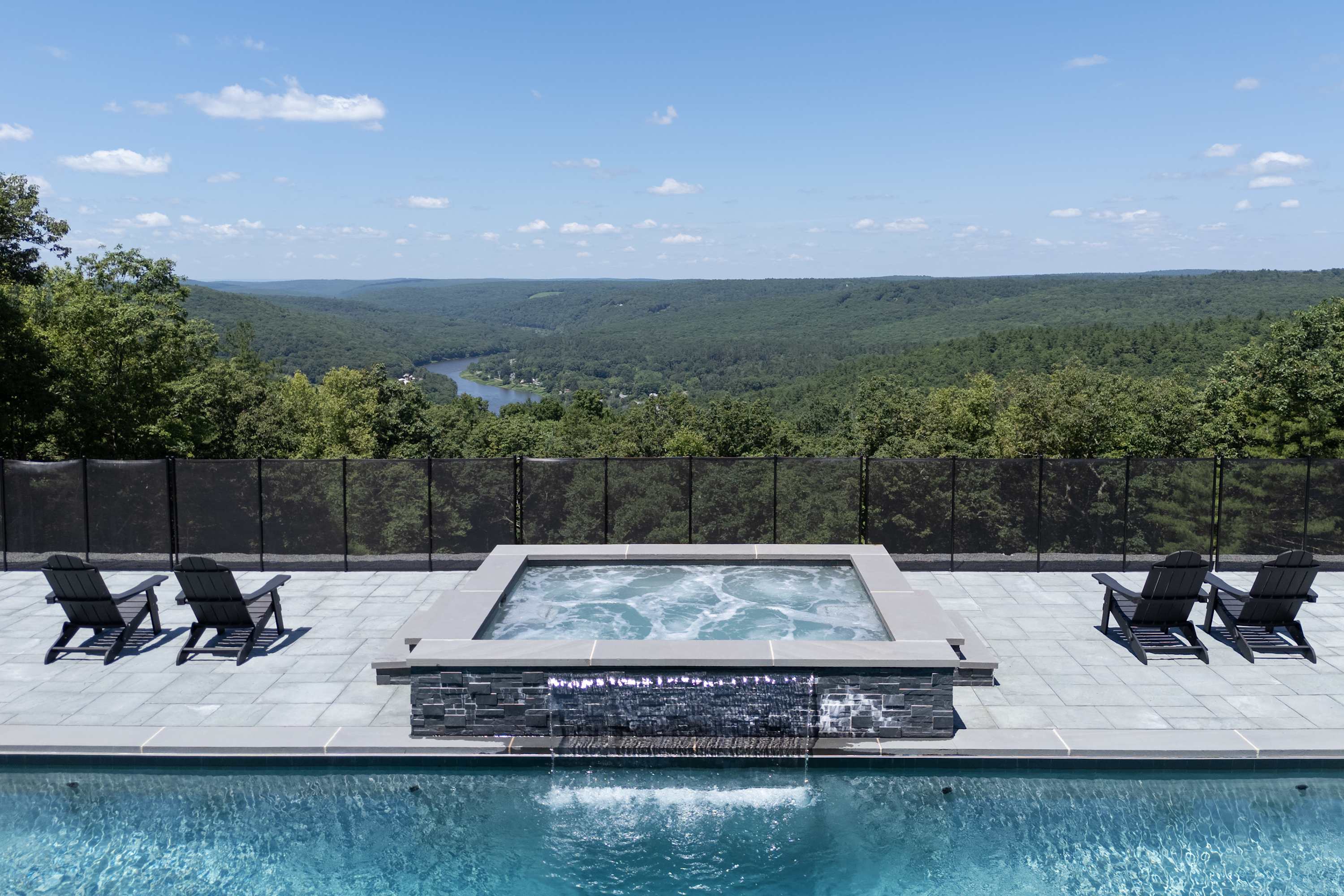 Outdoor hot tub with a waterfall feature flowing into a swimming pool, set against a stunning panoramic view of a lush green valley and river under a clear blue sky.