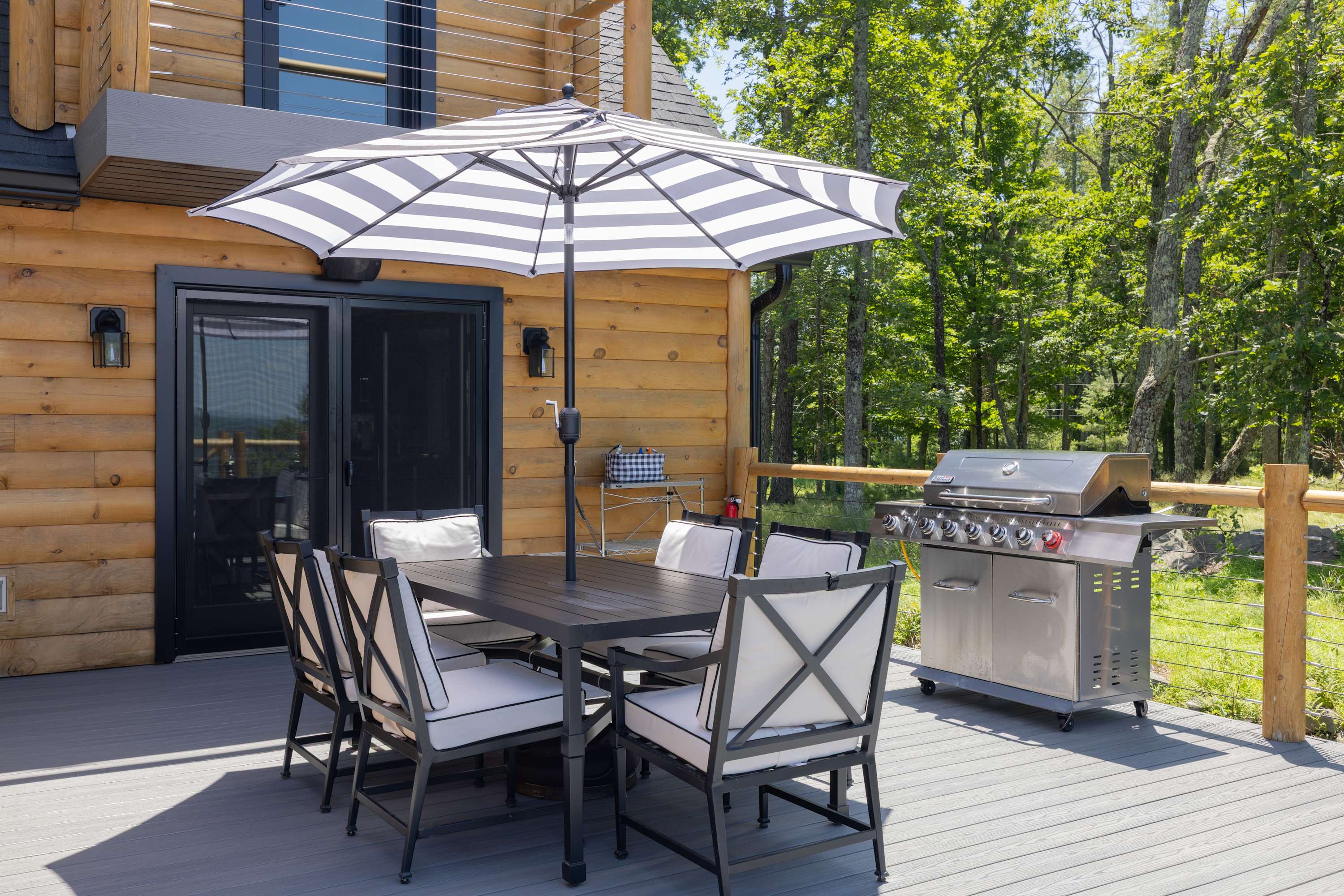 Outdoor dining area on a wooden deck, featuring a black dining table with six chairs and a striped umbrella, alongside a large stainless steel BBQ grill, surrounded by lush green trees.