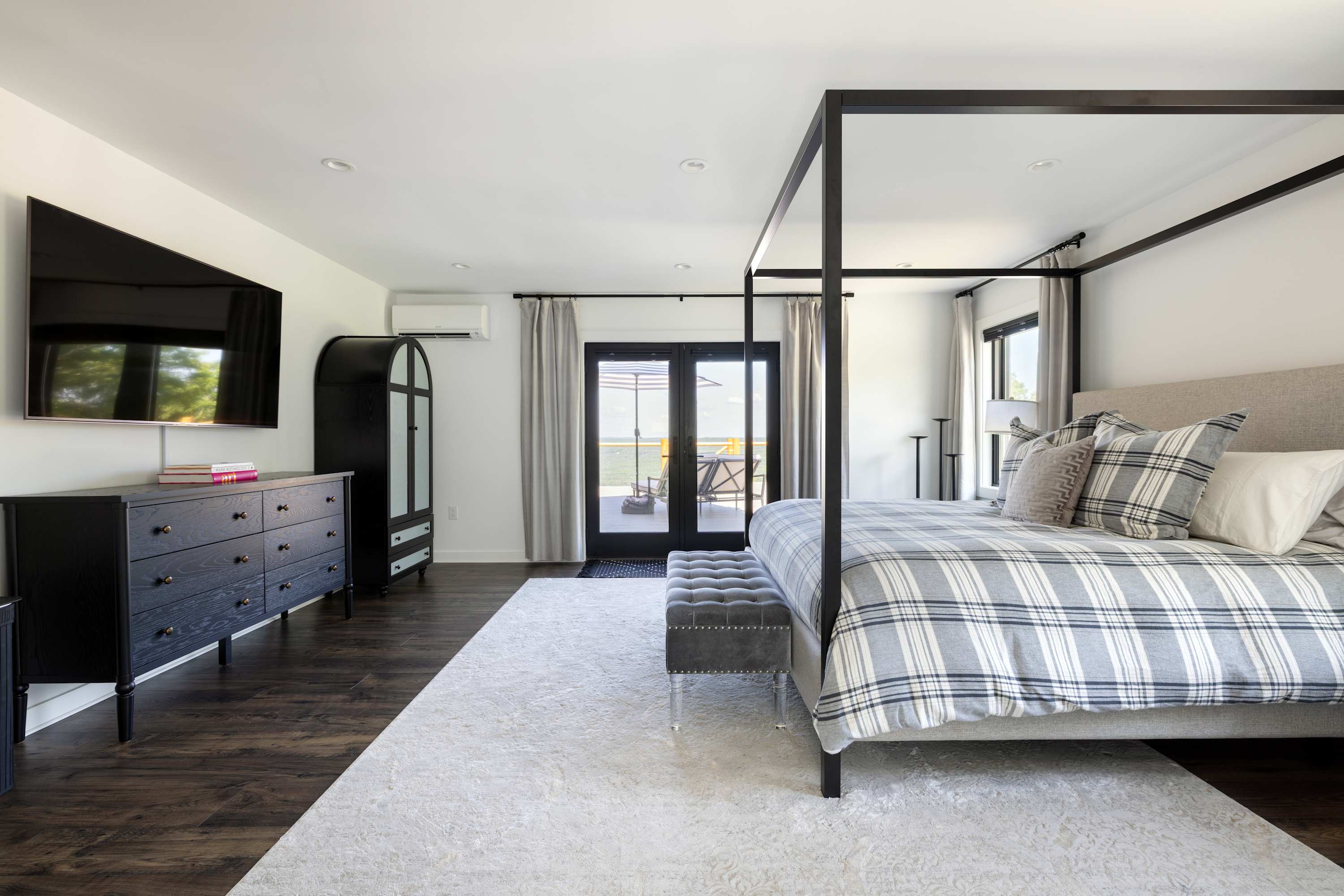 A spacious bedroom with a black four-poster king bed, dark hardwood floors, a large area rug, and a black dresser with a wall-mounted TV, offering access to an outdoor deck.