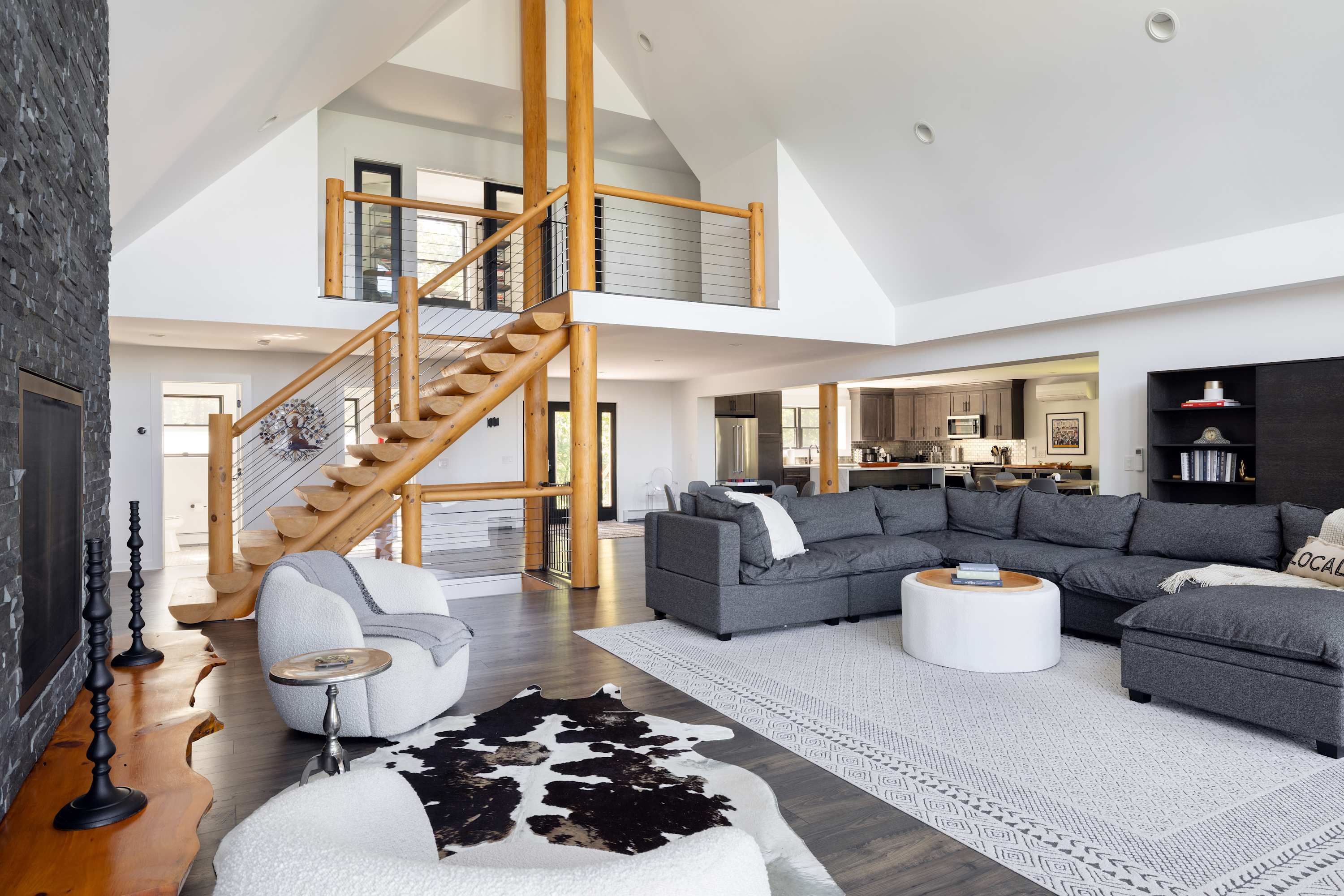 A grand living room with a large gray sectional sofa, a white ottoman, and a dark stone fireplace, featuring a unique wooden staircase with cable railings and a cowhide rug.